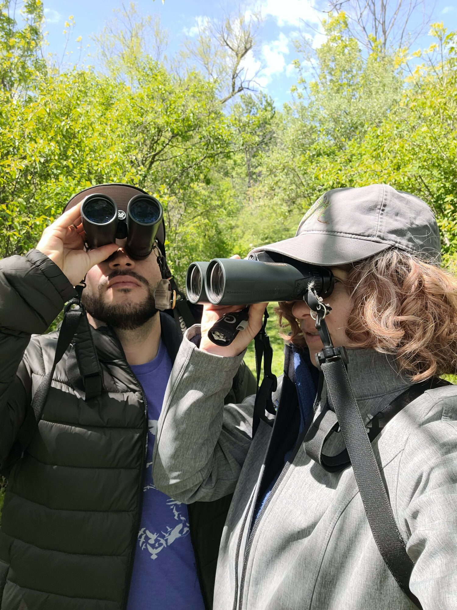 Two people outdoors, one holding binoculars and the other using a camera, surrounded by green trees and blue sky.