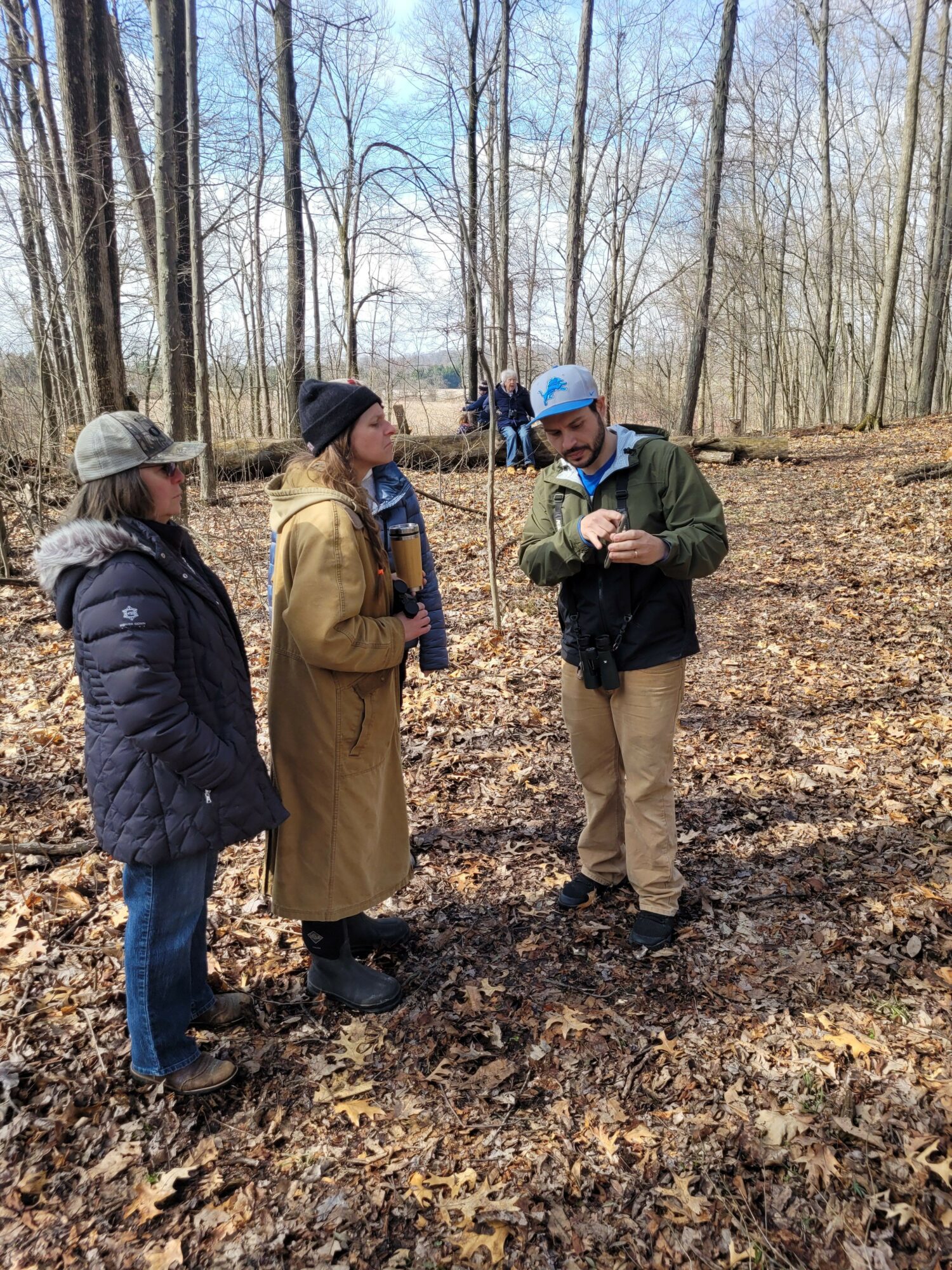 Two people standing in a leaf-covered forest, talking, with trees and a fence in the background.
