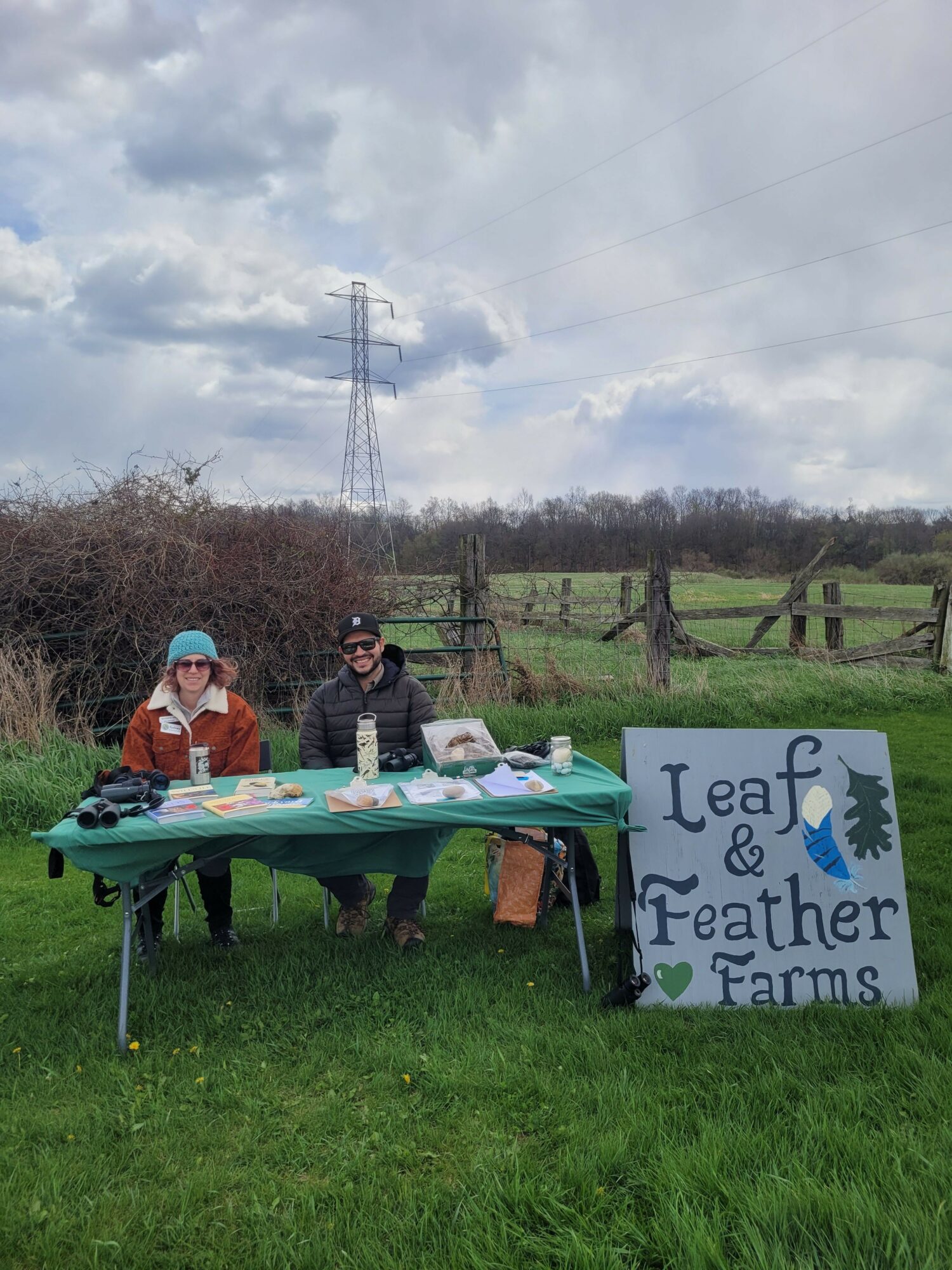 Two people sit at a table outdoors near a sign that reads 'Leaf & Feather Farms,' with green grass and cloudy sky in background.
