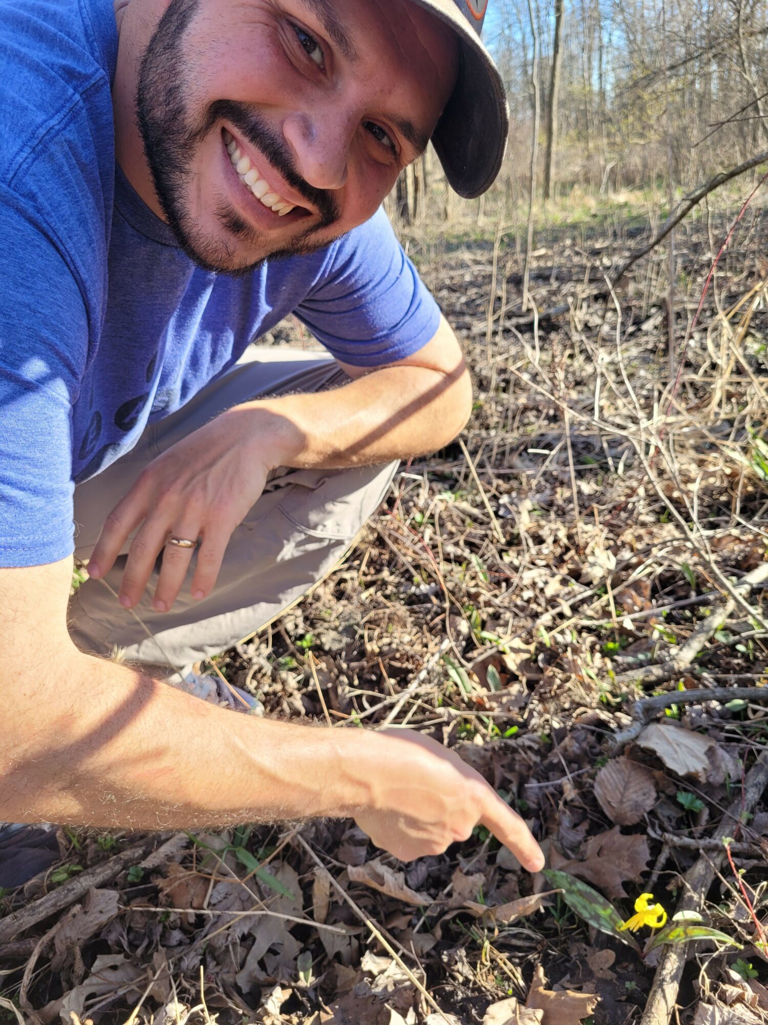 Smiling man outdoors pointing at a small yellow duck figurine on the ground, surrounded by dry leaves and twigs.