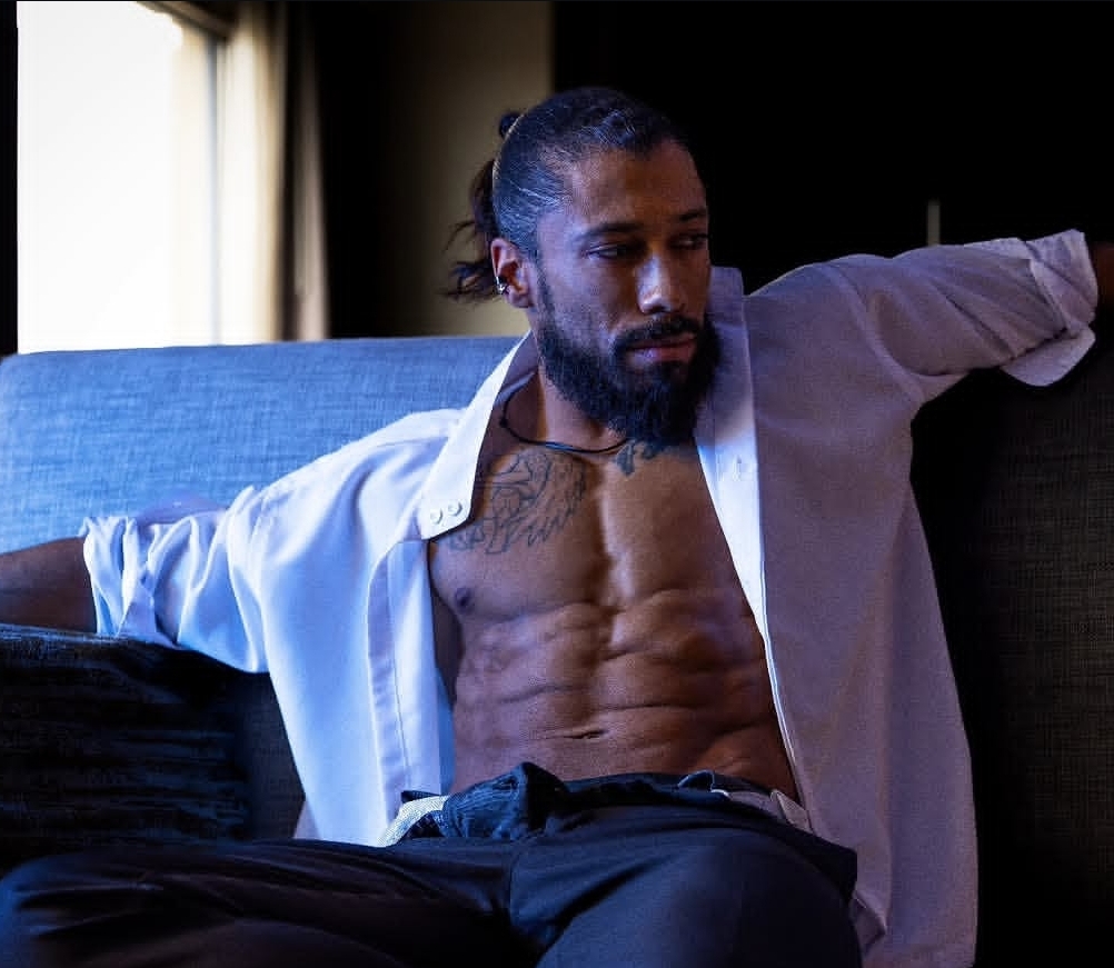 Man with a beard and long hair, shirt open, sitting on a couch in a dimly lit room.