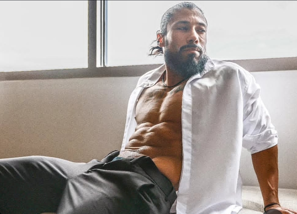 Man with a beard and long hair, shirt open, sitting on a window ledge, showing his chest and abs.