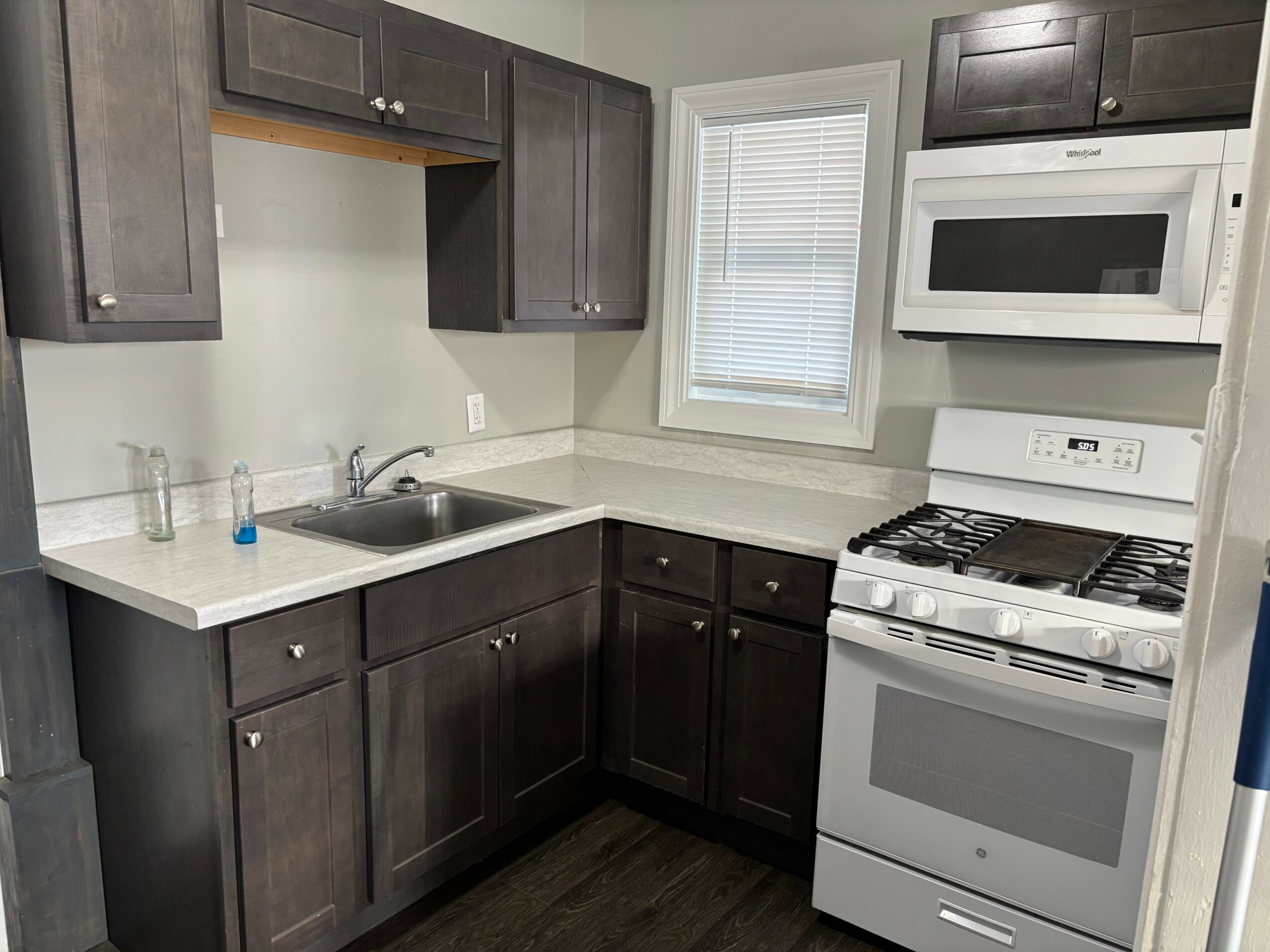 Kitchen with dark cabinets, white countertop, stainless steel sink, microwave, and stove, window above the countertop, and light-colored walls.