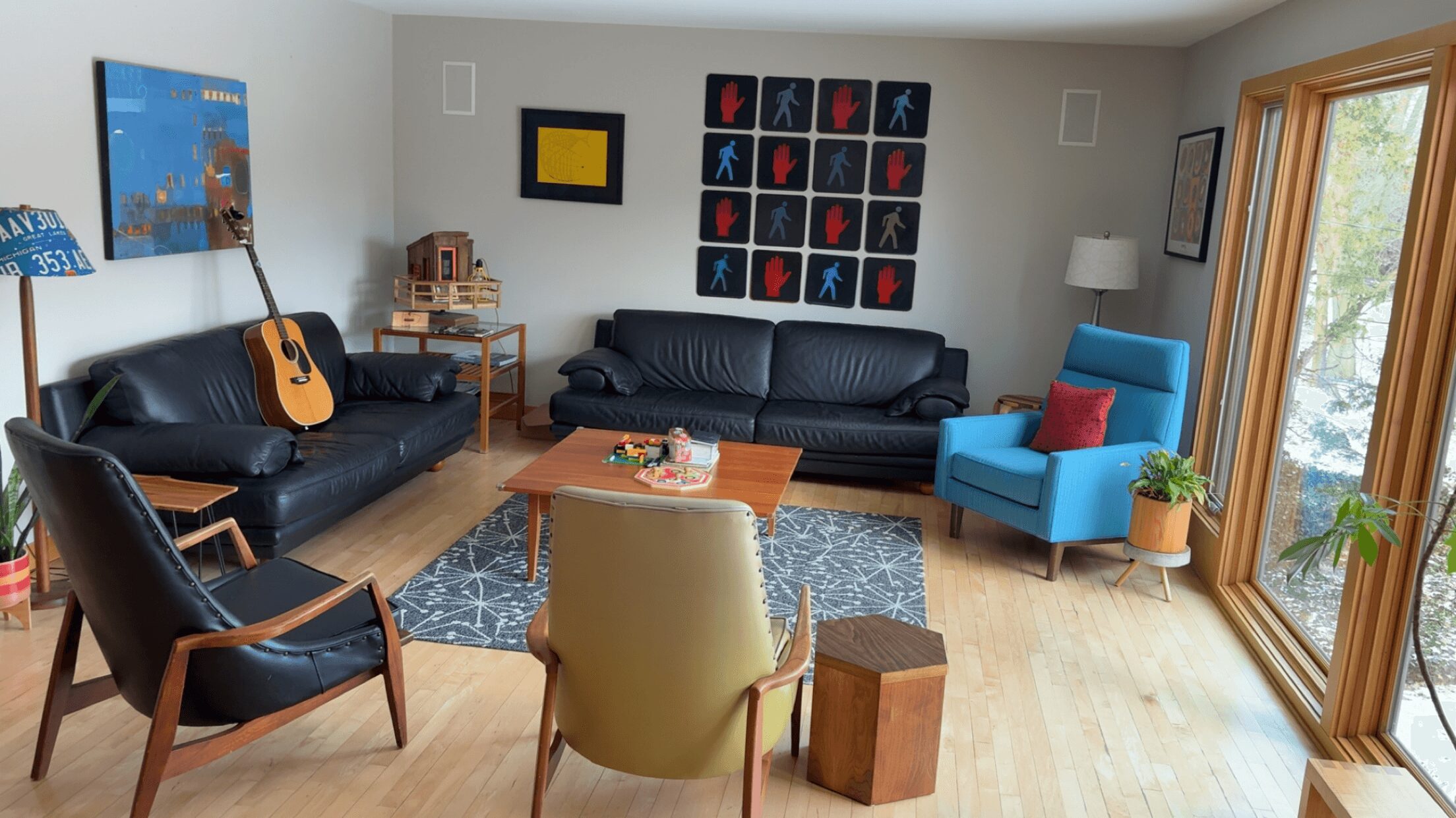 Living room with black sofas, a blue armchair, a wooden coffee table, and wall art, with large windows showing trees outside.