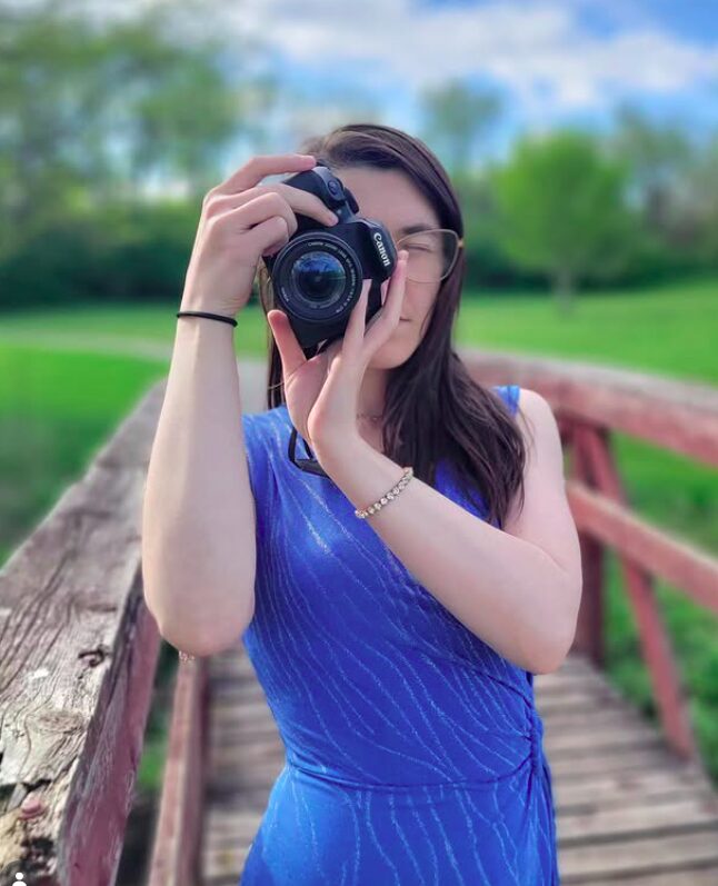 Person taking a photo with a camera outdoors on a wooden bridge, wearing a blue dress, with trees and sky in background.