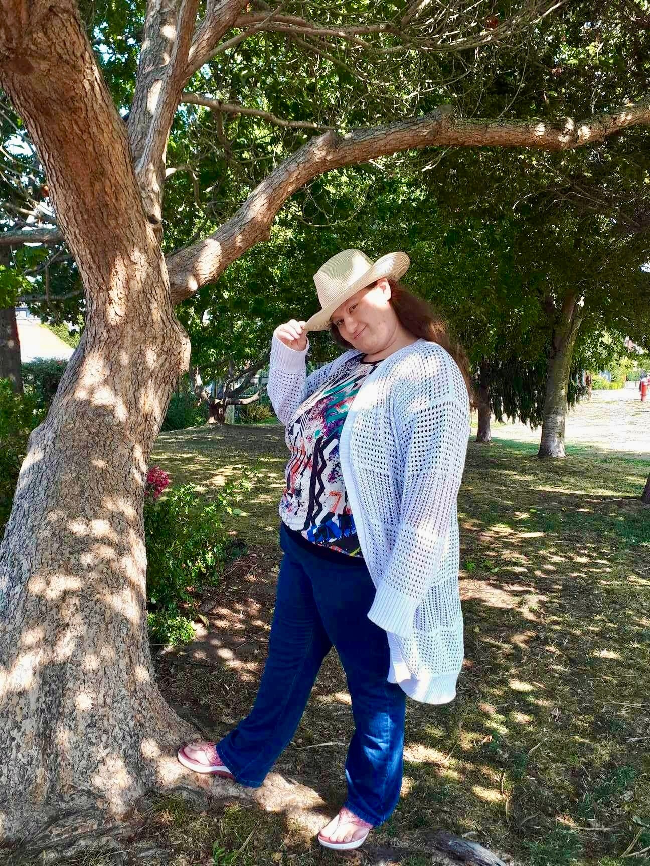 Woman wearing a hat, standing next to a tree in a park with green trees and grass.