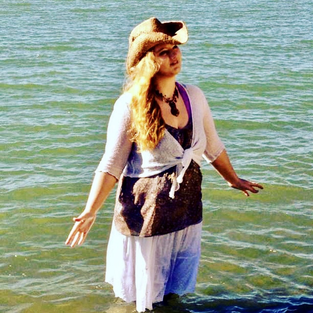 Woman standing in water at the beach, wearing a hat, sunglasses, and a dress, with arms outstretched.