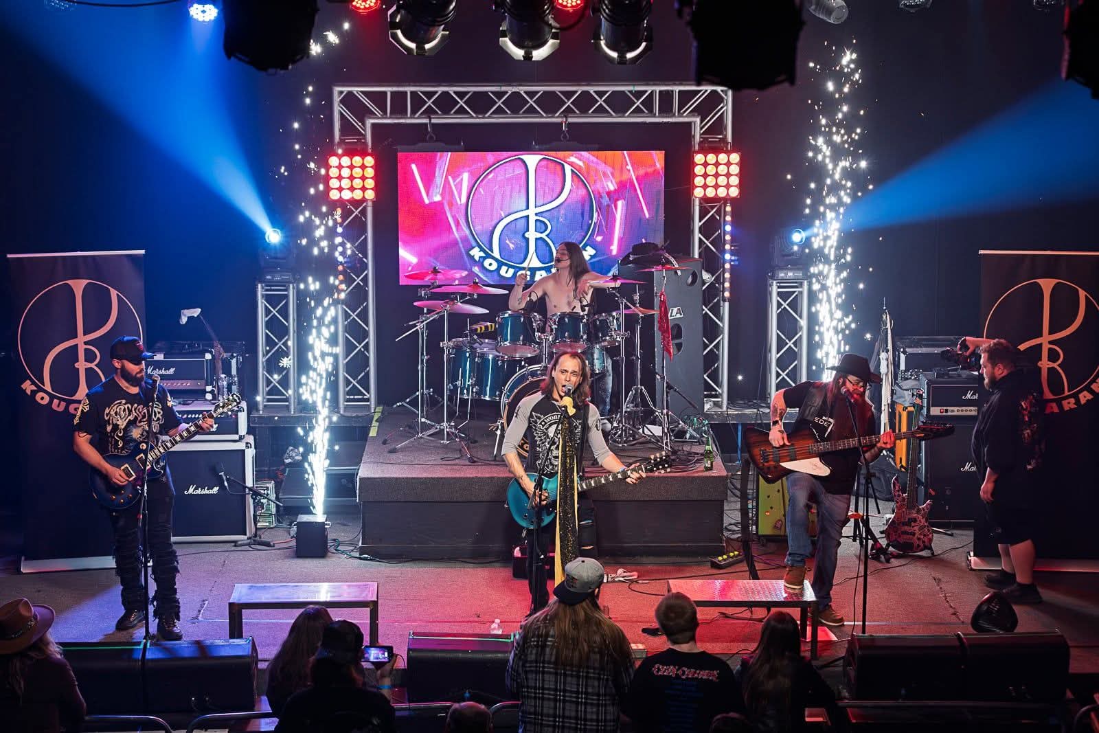 Band performs on stage with colorful lights and fireworks, audience watching in front, large screen with logo behind the band.