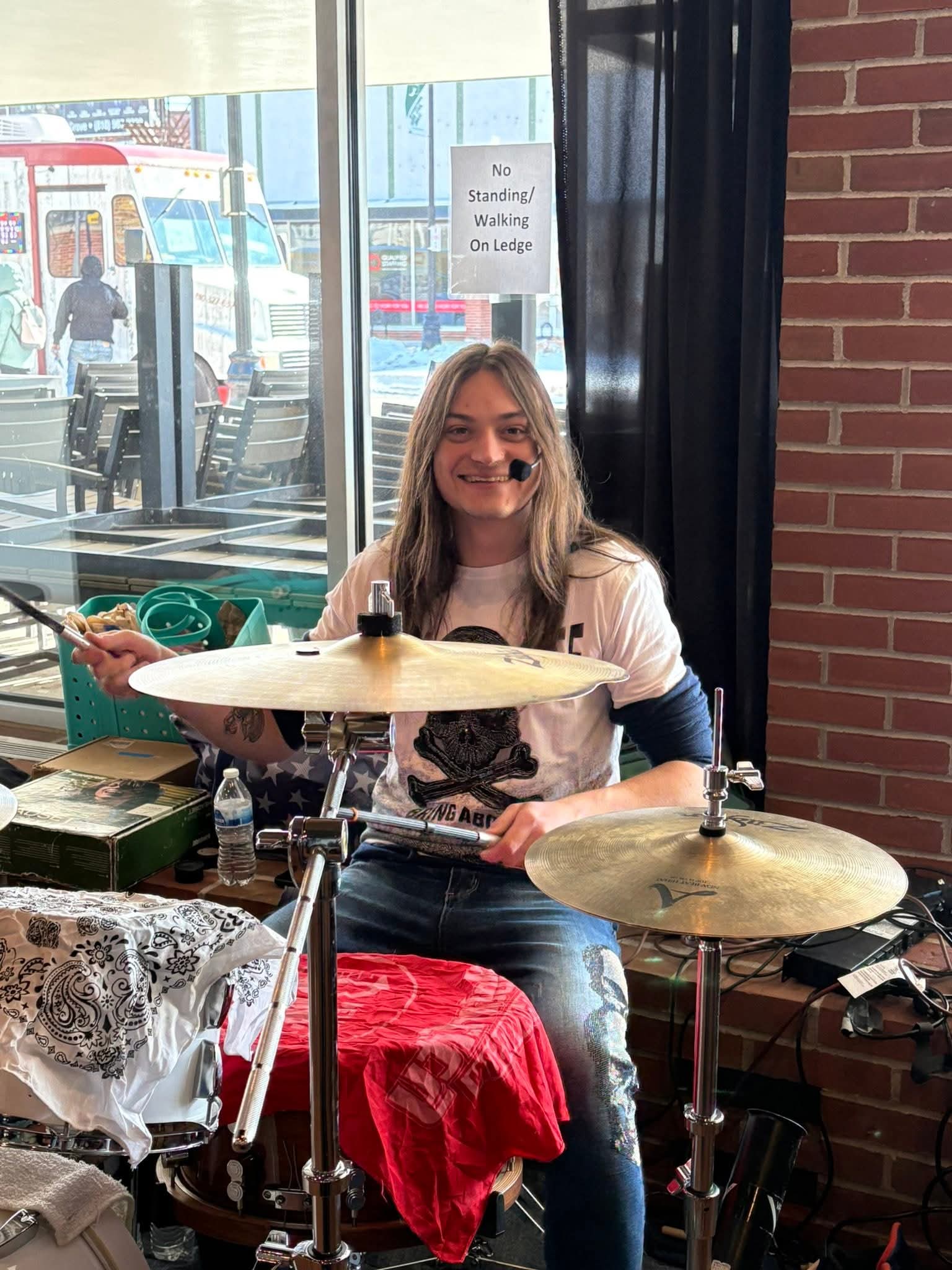 Young woman smiling, sitting at a drum set inside a cafe, with a window and brick wall behind her.