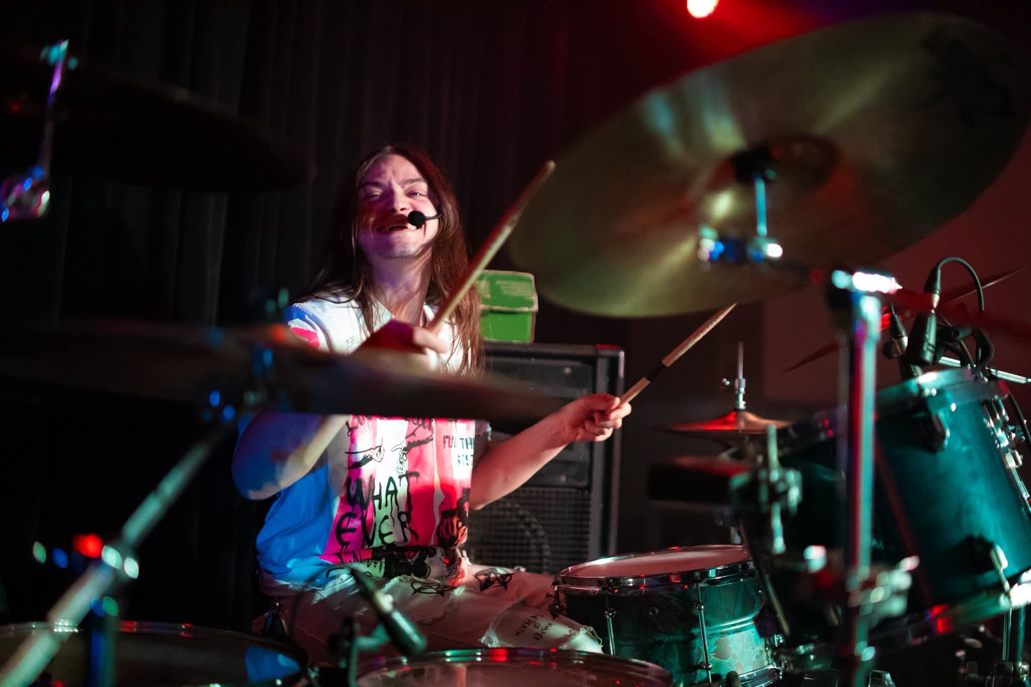 A person playing drums on stage, smiling, with a dark background and stage lighting, wearing a colorful shirt.
