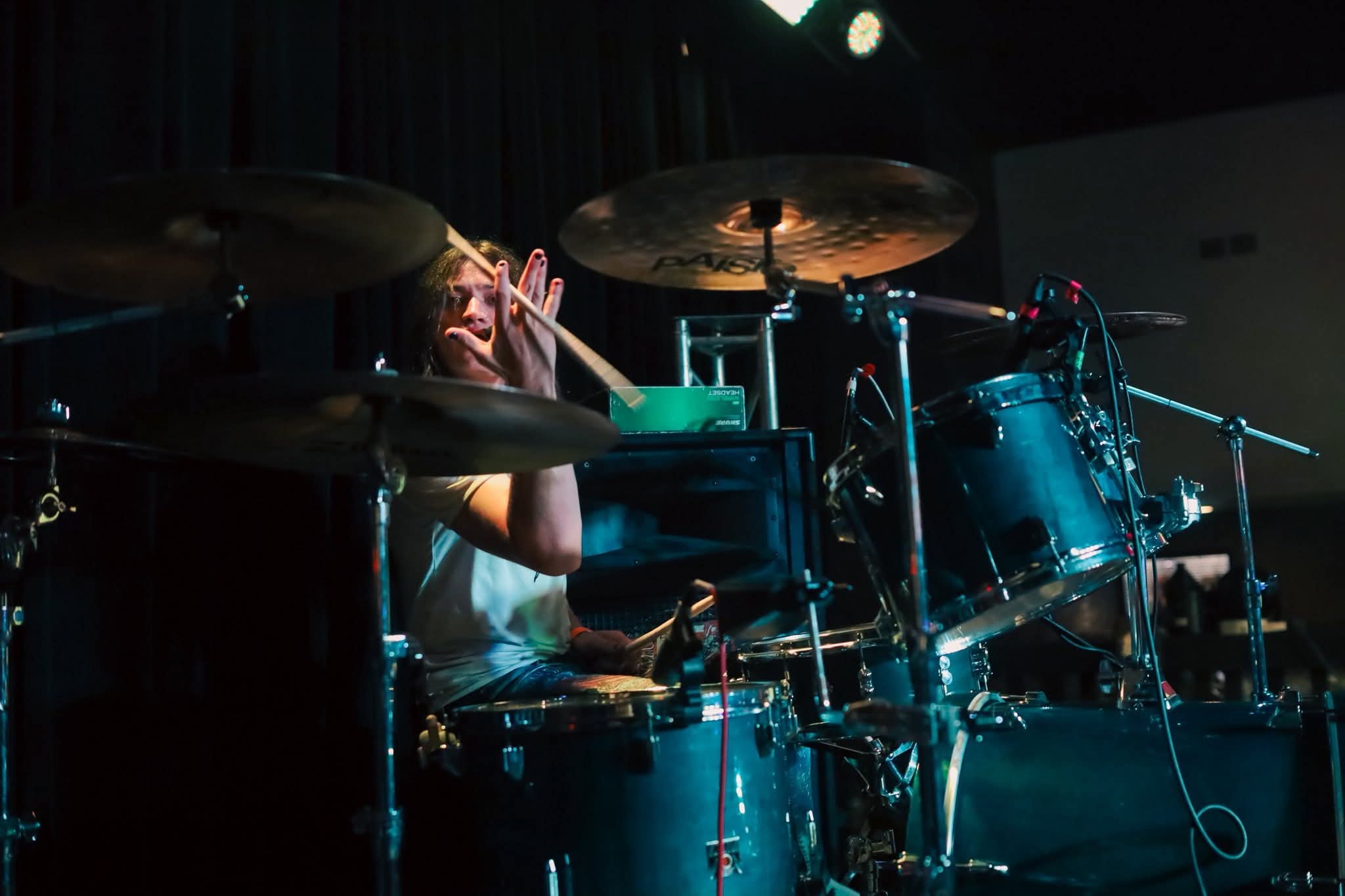 Person playing drums on stage with cymbals and drum set, dark background, stage lighting.