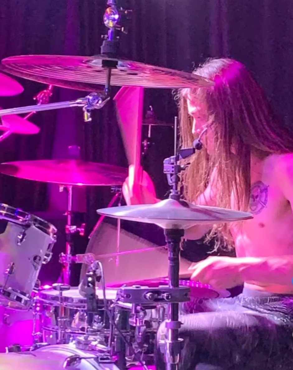 Person playing drums on stage with purple lighting, long hair, and a tattoo on their arm.