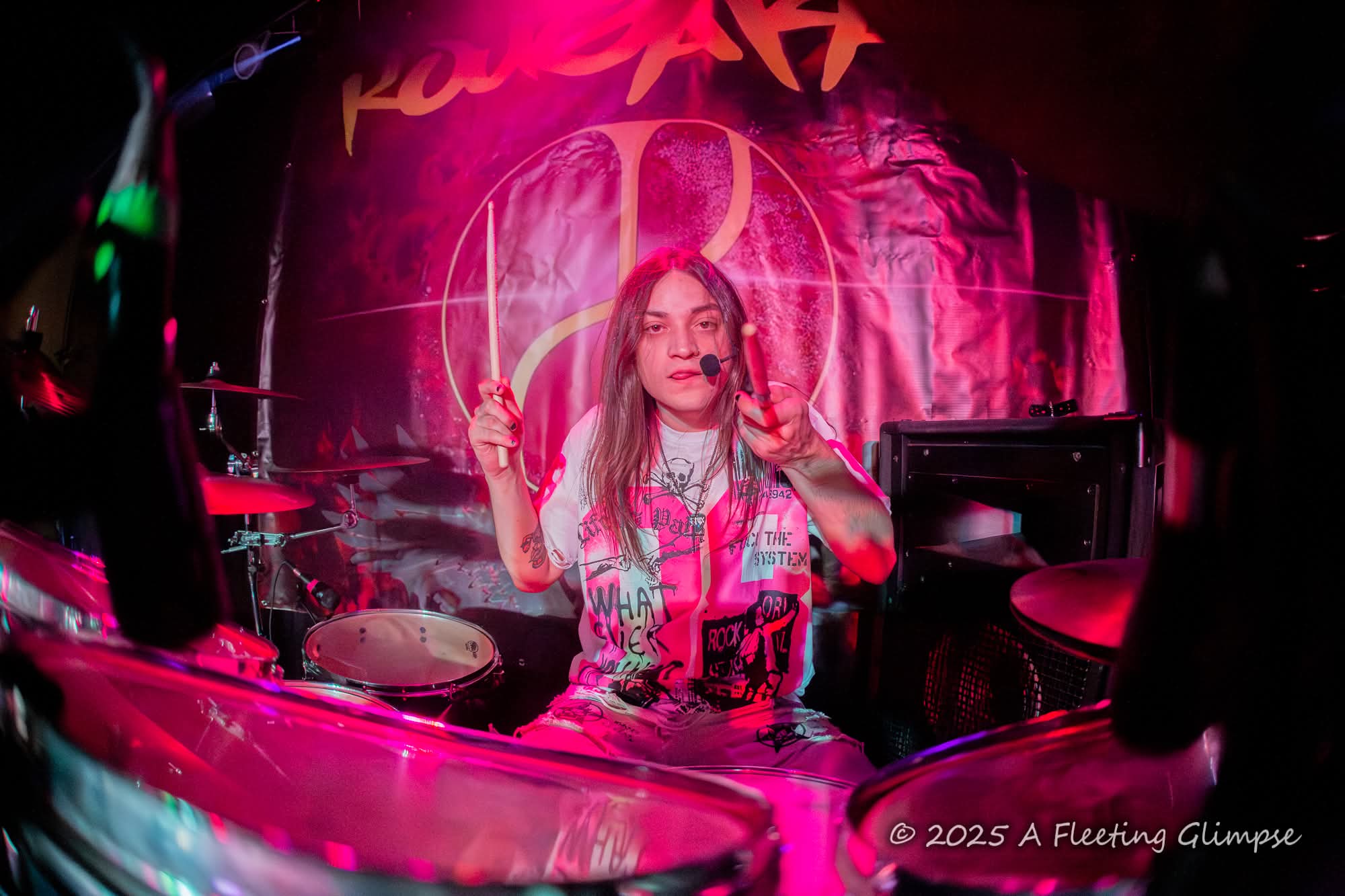 Female musician performing on stage with pink lighting and a drum set in the background.