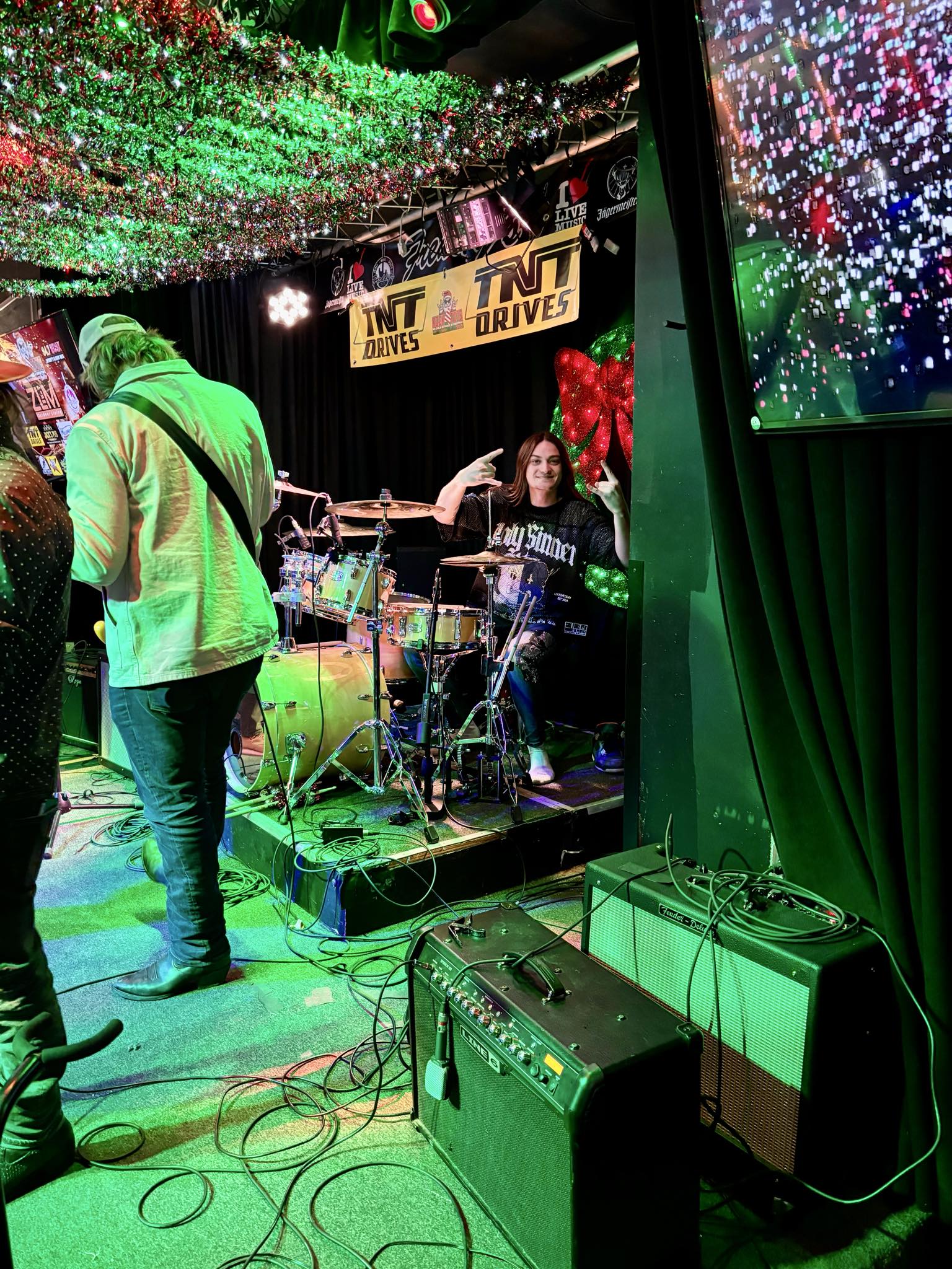 Musicians perform on stage with colorful lights and a sign reading 'LUV DRIVES,' surrounded by equipment and audience.
