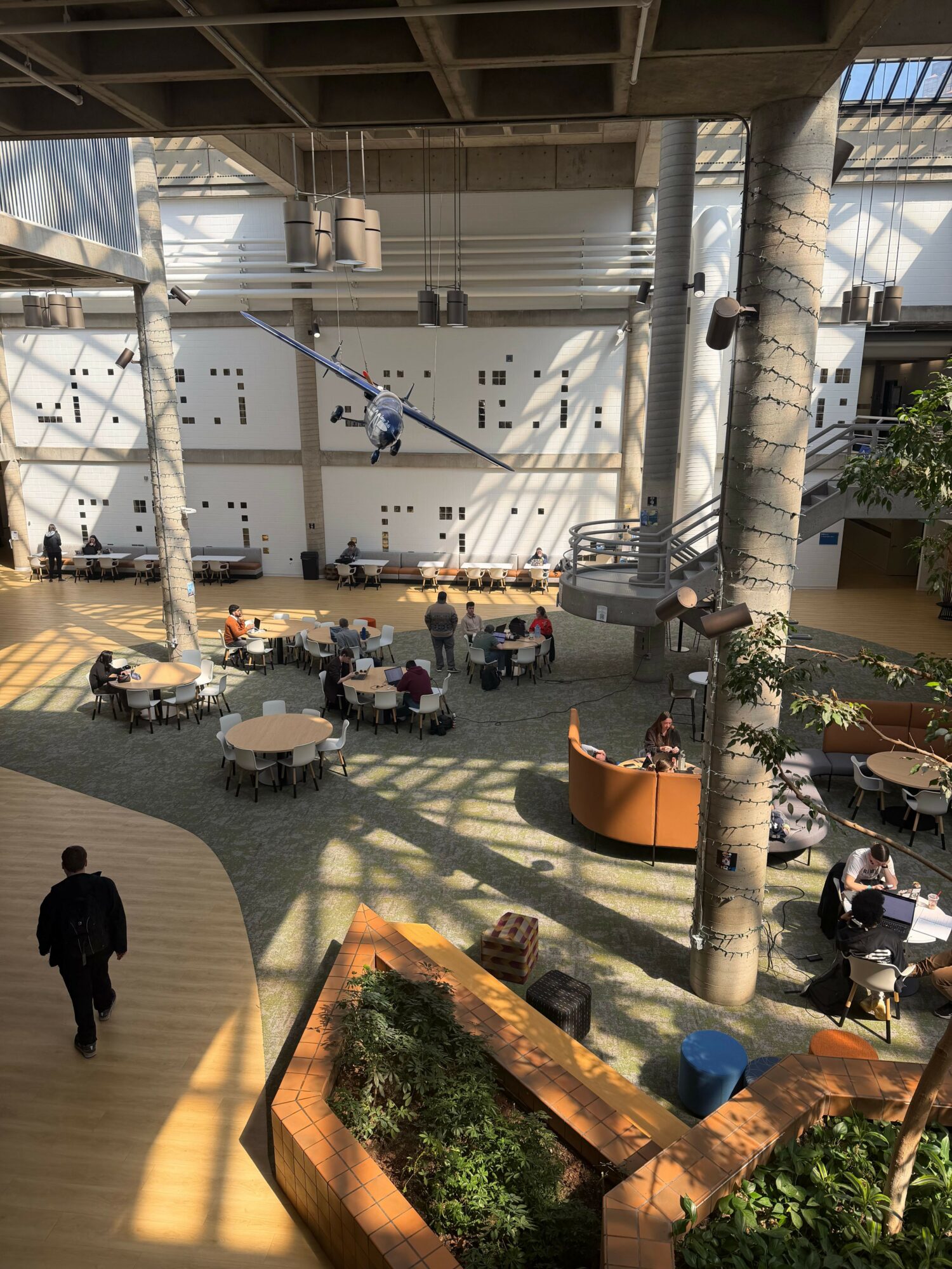 People gather in an outdoor courtyard with trees, tables, and chairs, surrounded by buildings and sunlight casting shadows.