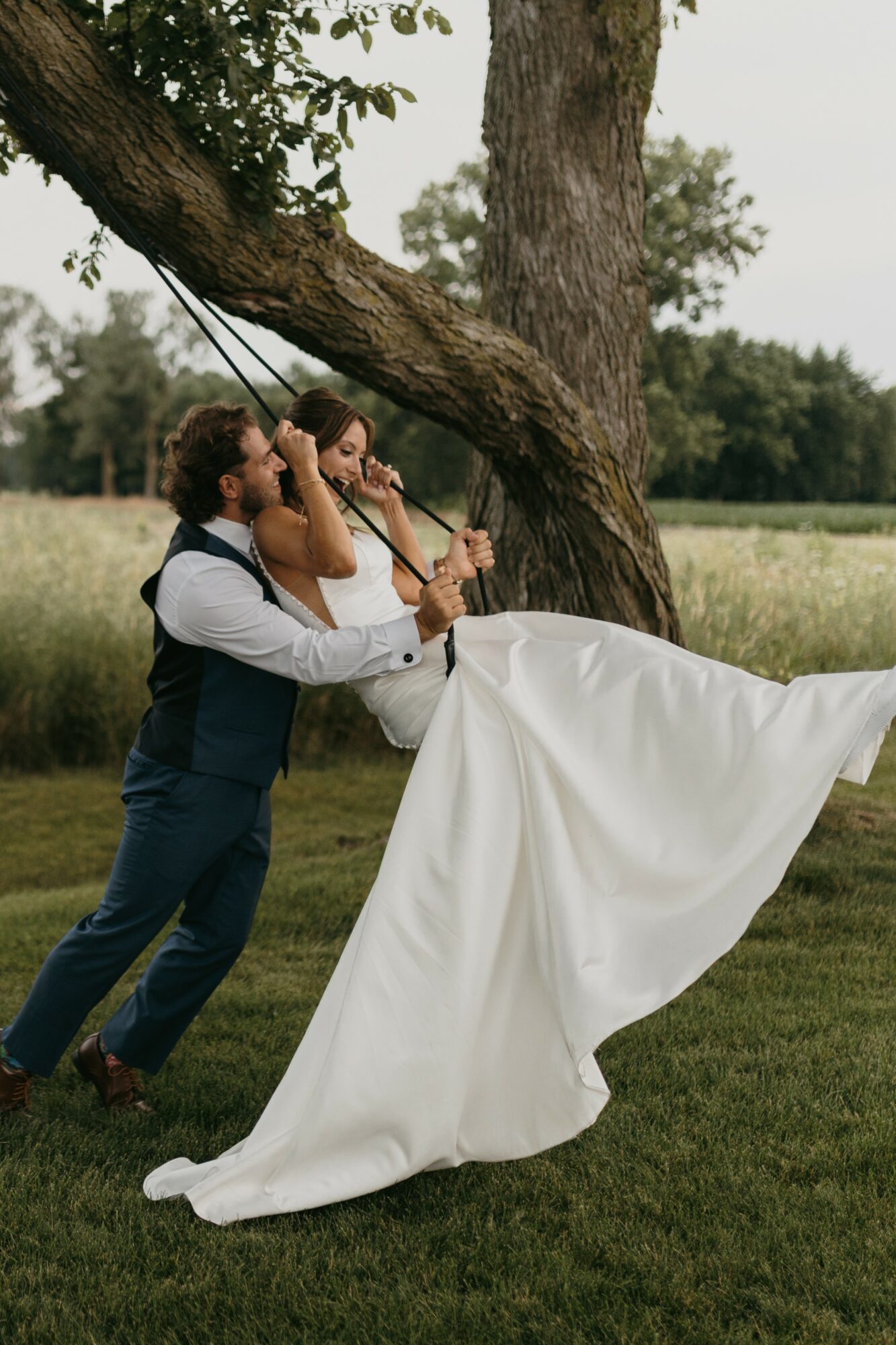 Two people in formal attire on a swing hanging from a tree, with a grassy field in the background.