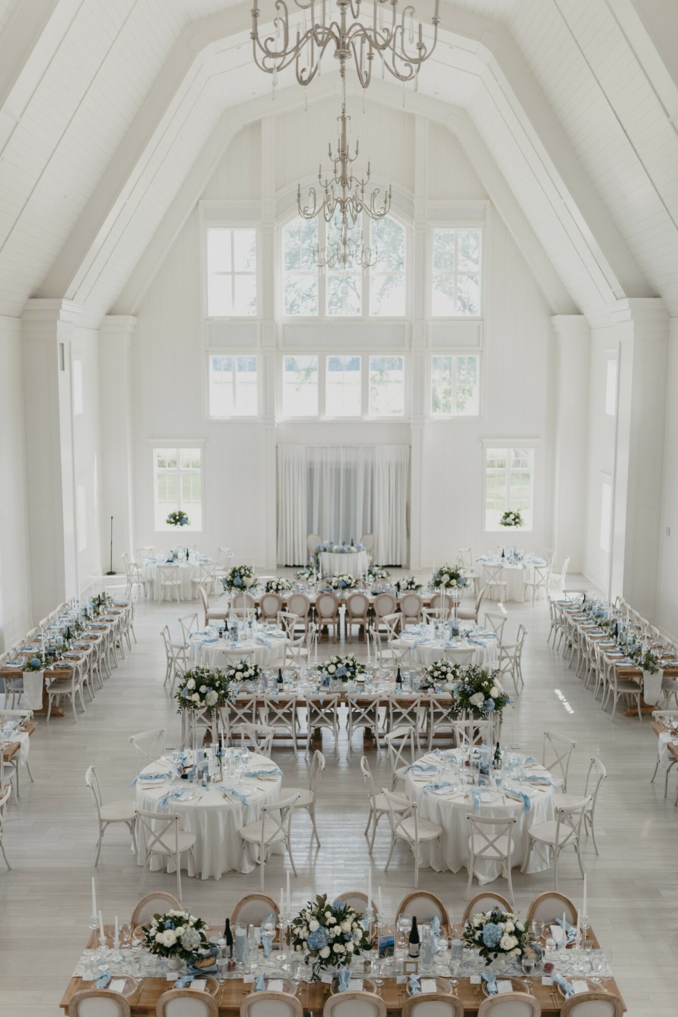 Elegant wedding reception hall with high vaulted ceiling, large windows, round tables with white tablecloths, floral centerpieces, and chandeliers.