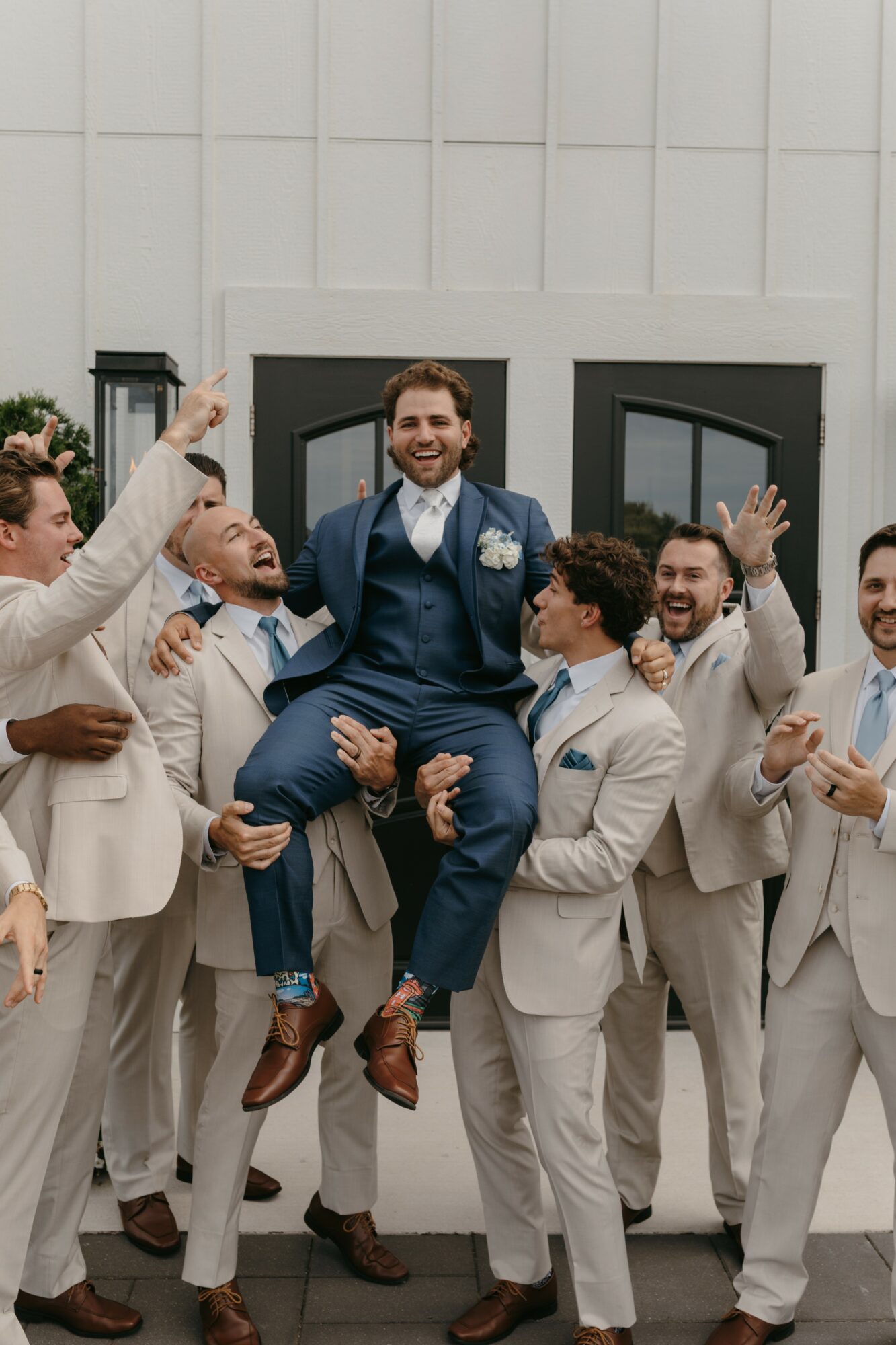Group of men in beige suits lifting a smiling man in a blue suit outdoors, celebrating.