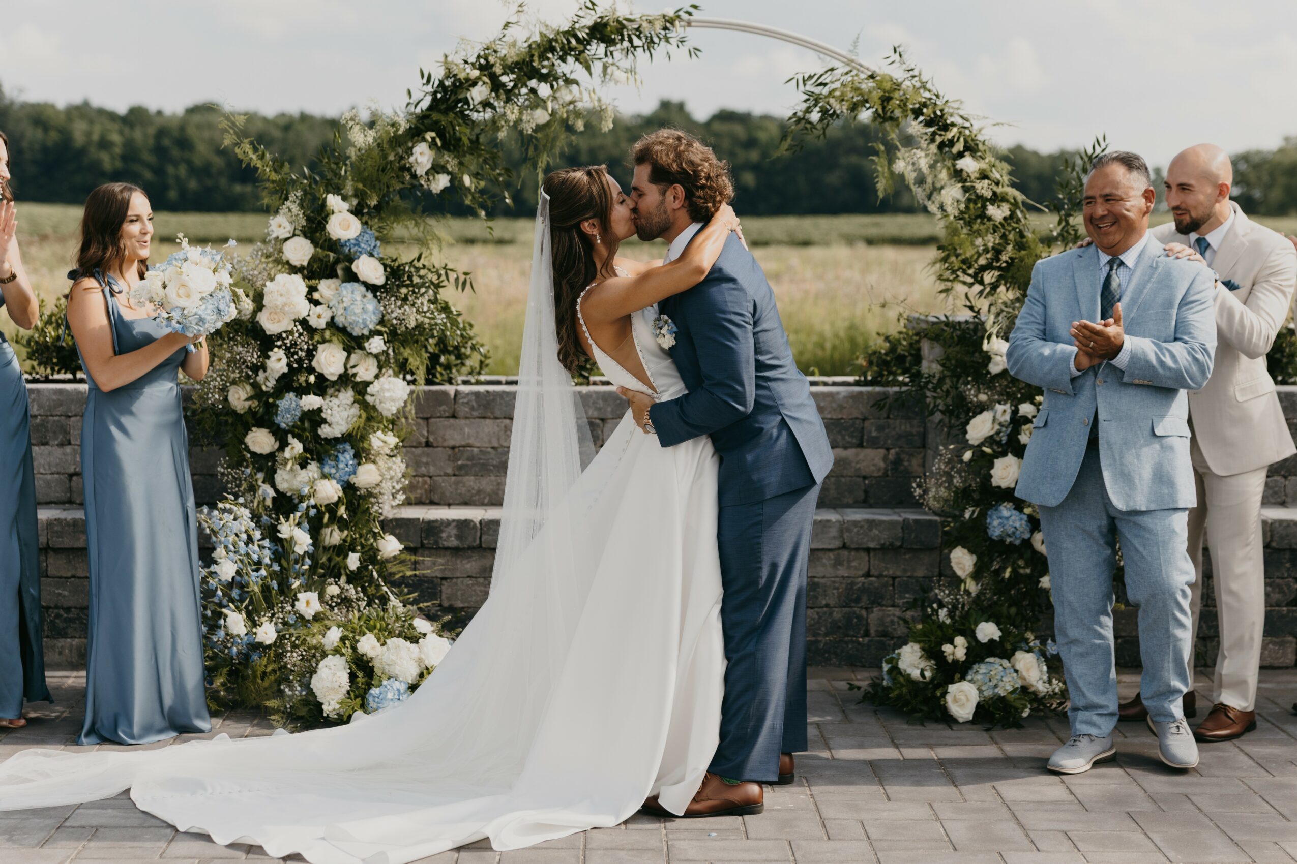 Couple kissing at outdoor wedding ceremony with floral arch, bridesmaids, and groomsmen, on a sunny day.