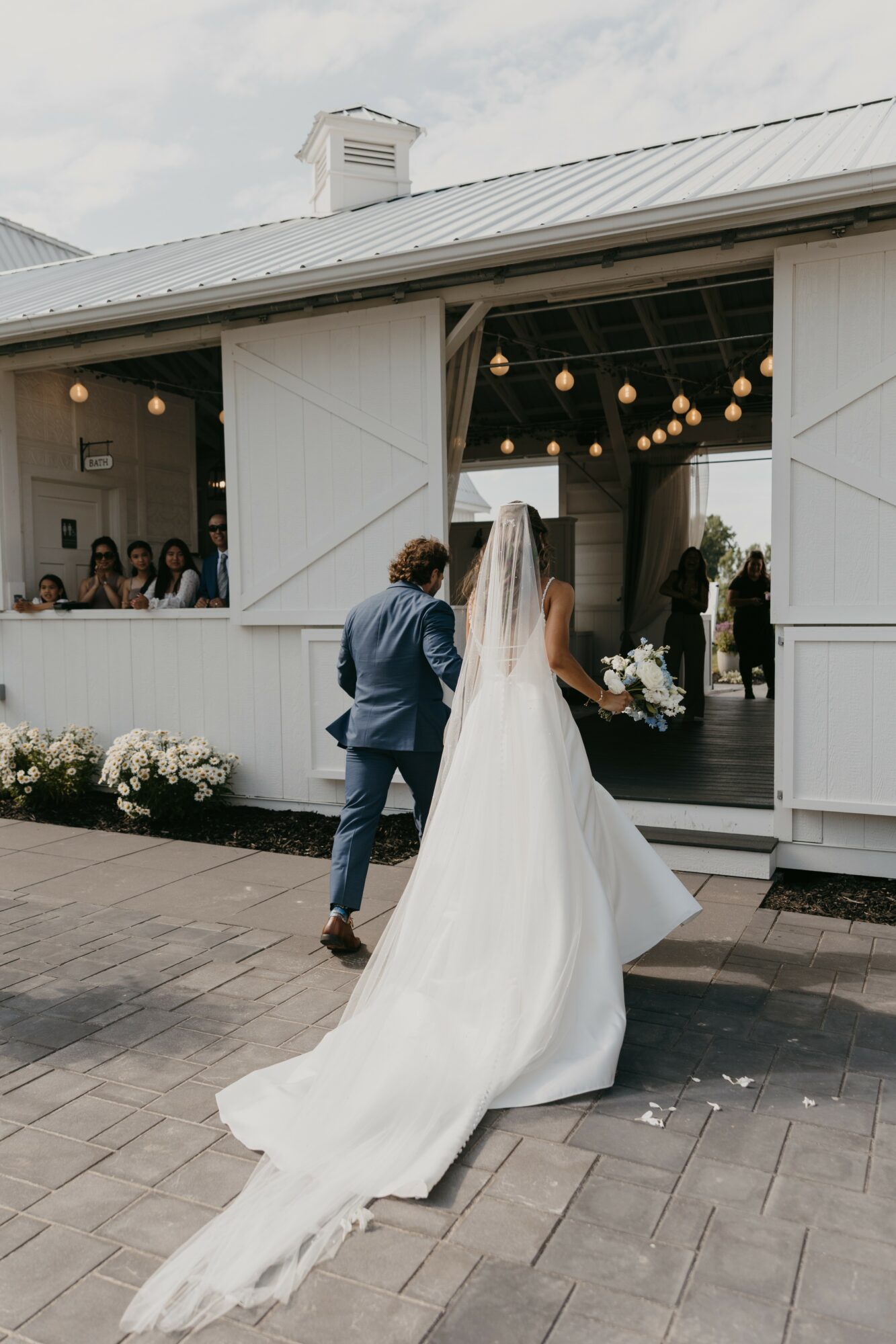 Bride and groom walking out of a white building with open doors, wedding guests inside, bride holding bouquet, bride's train flowing behind.
