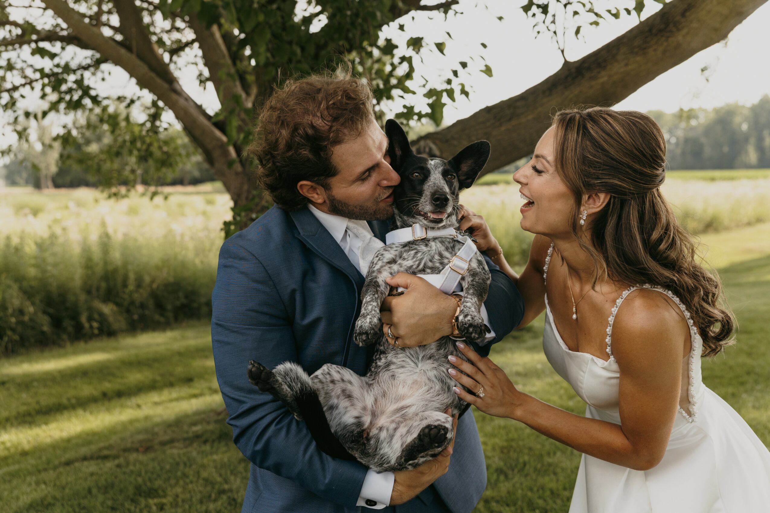 A couple with a dog outdoors, the man holding the dog and the woman touching its face, under a tree.