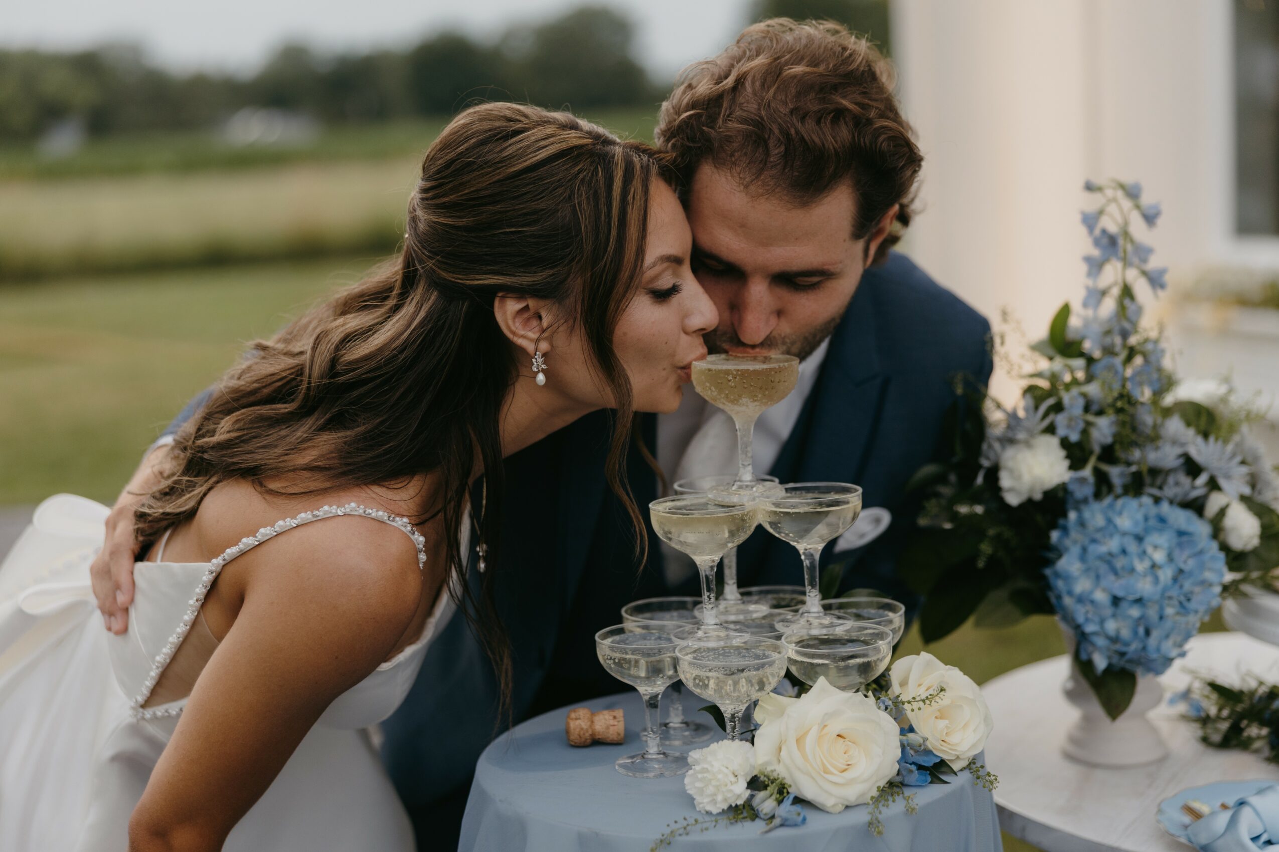 A bride and groom share a toast at their wedding, leaning close with champagne glasses, outdoors with flowers nearby.