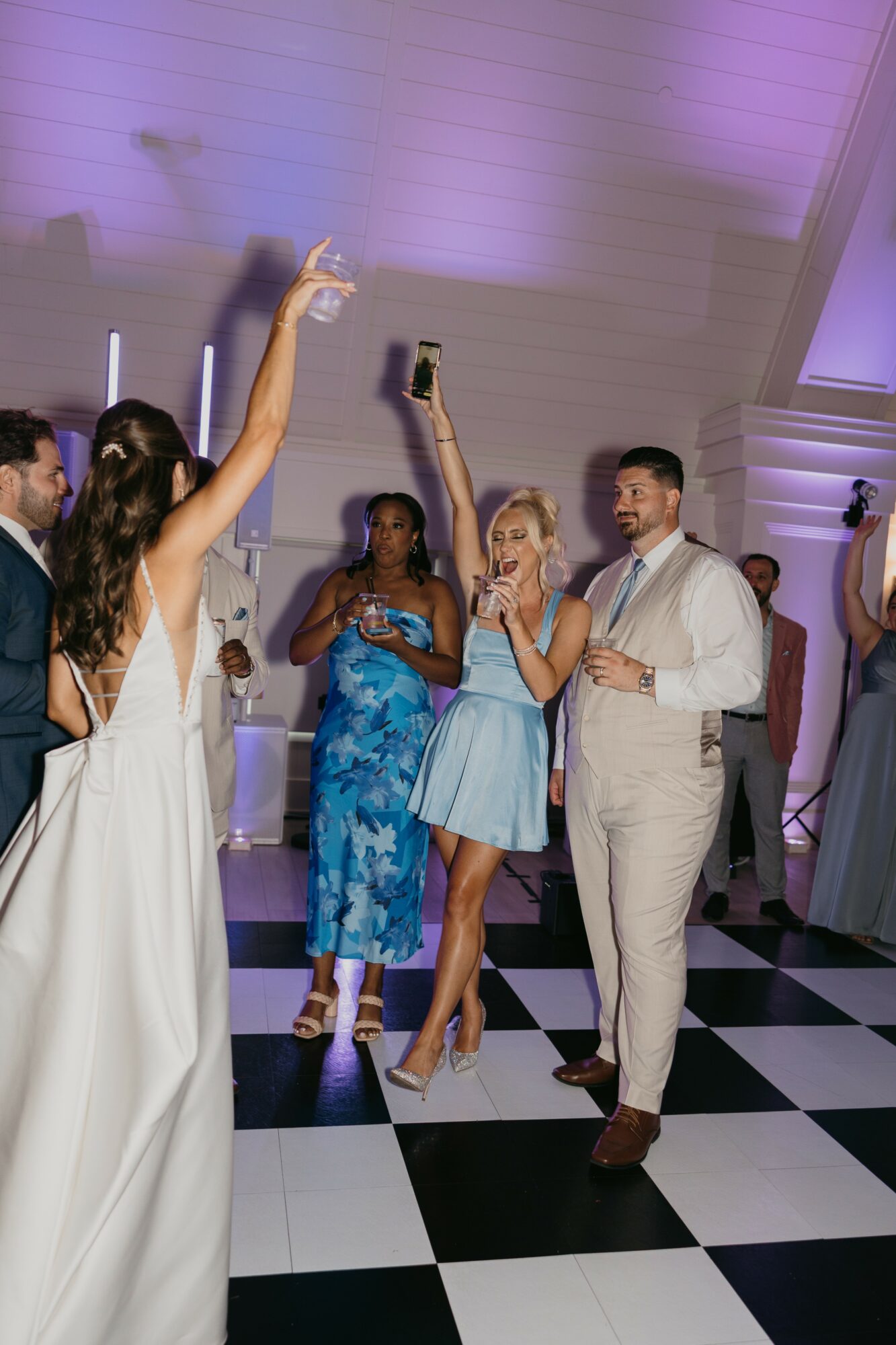 Group of people dancing and taking photos at a celebration, with a checkered floor and purple lighting, in formal attire.