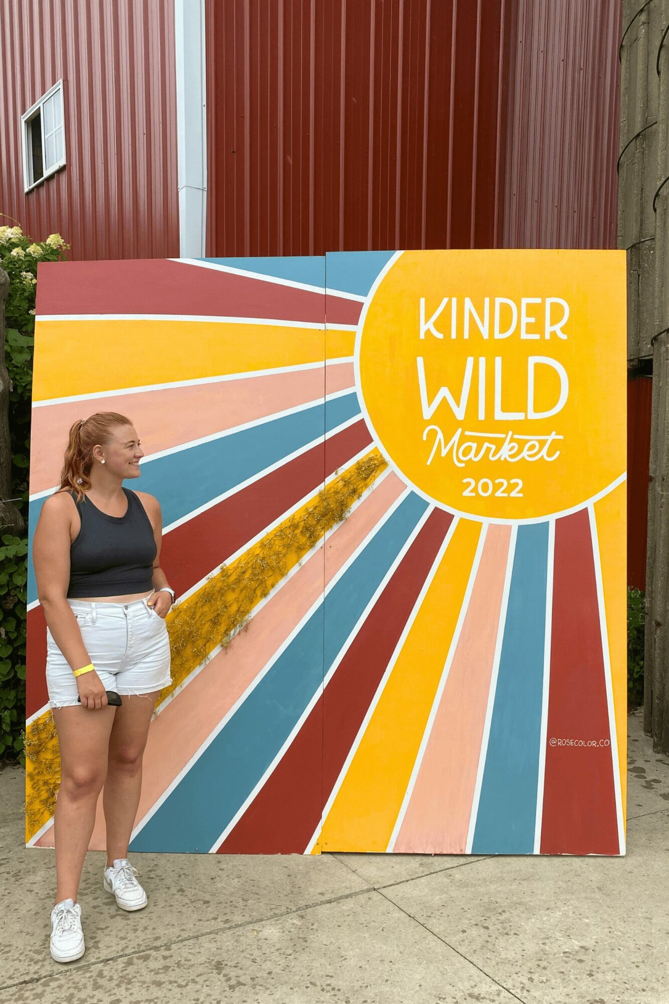 Young woman standing next to a colorful sign with the text 'KINDER WILD Market 2022' and a rainbow design.