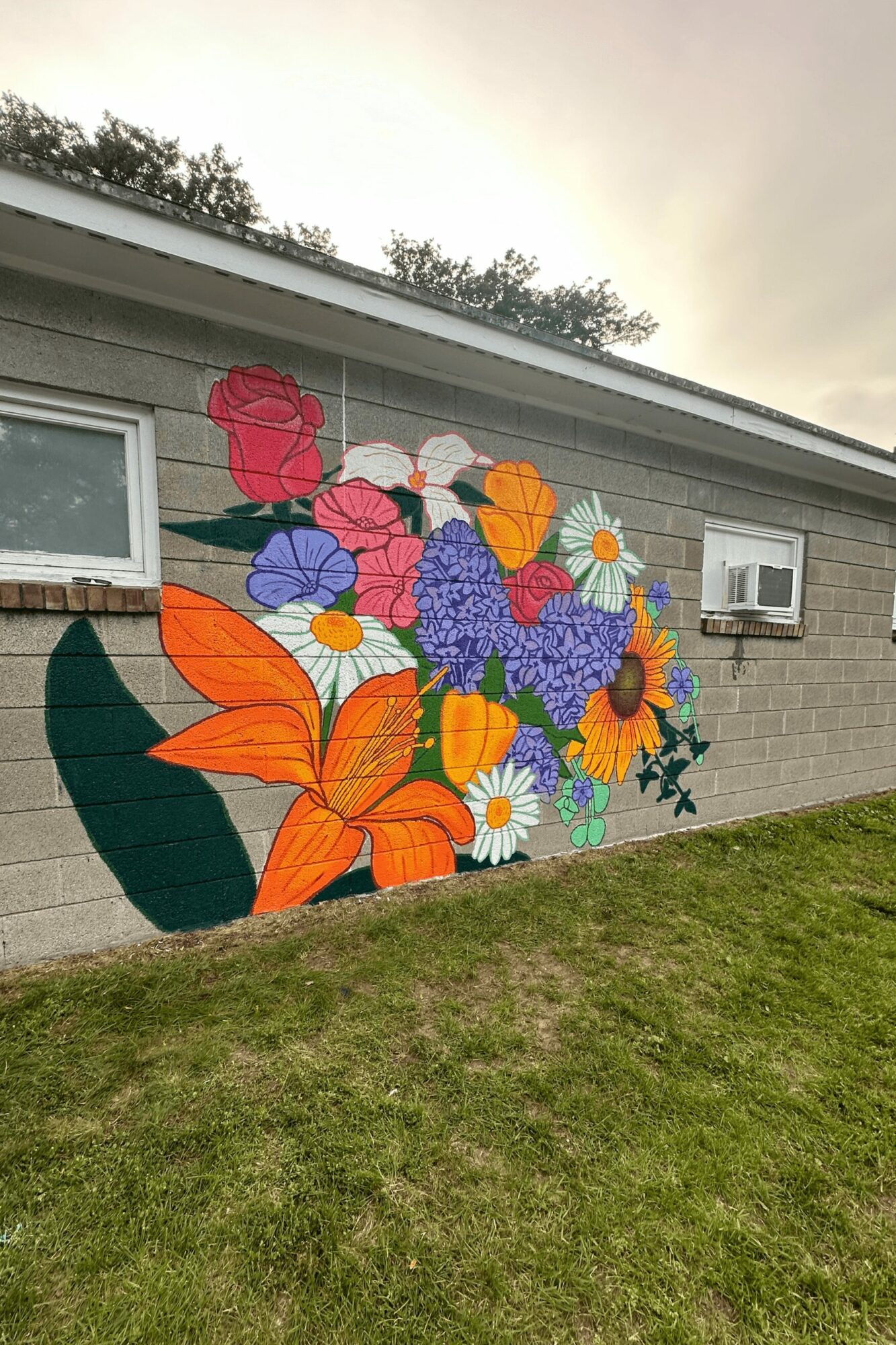 Colorful floral mural on brick wall with various flowers and leaves, window, and air conditioning unit, grass in foreground.