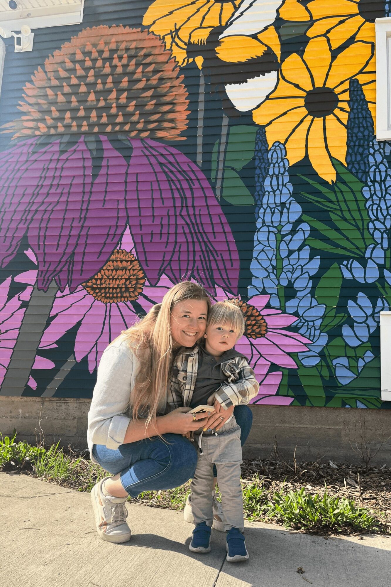 Woman crouching with a child in front of a colorful floral mural, smiling outdoors.