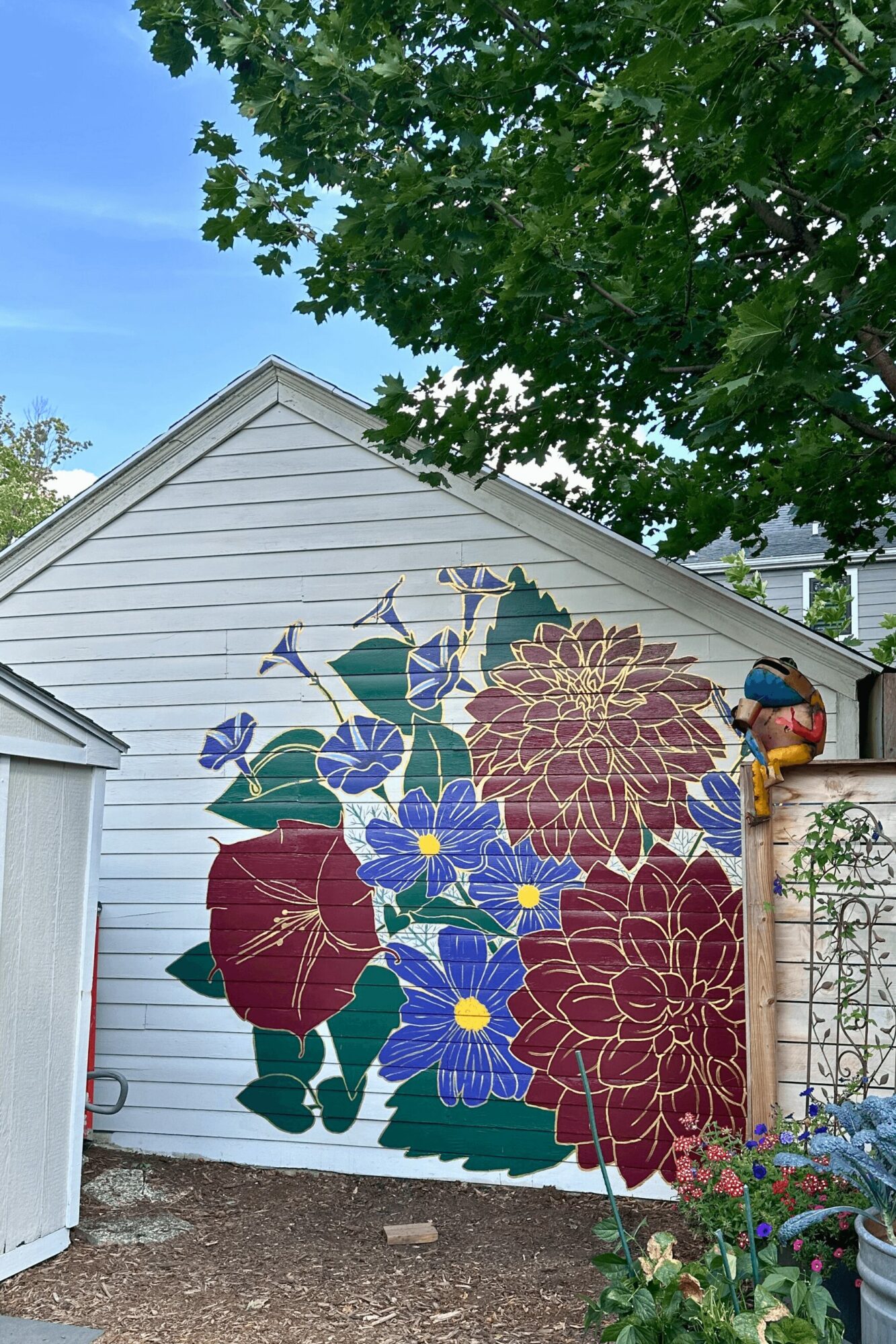 Garage door with a large floral mural featuring red, purple, and blue flowers and green leaves, under a tree with green foliage.