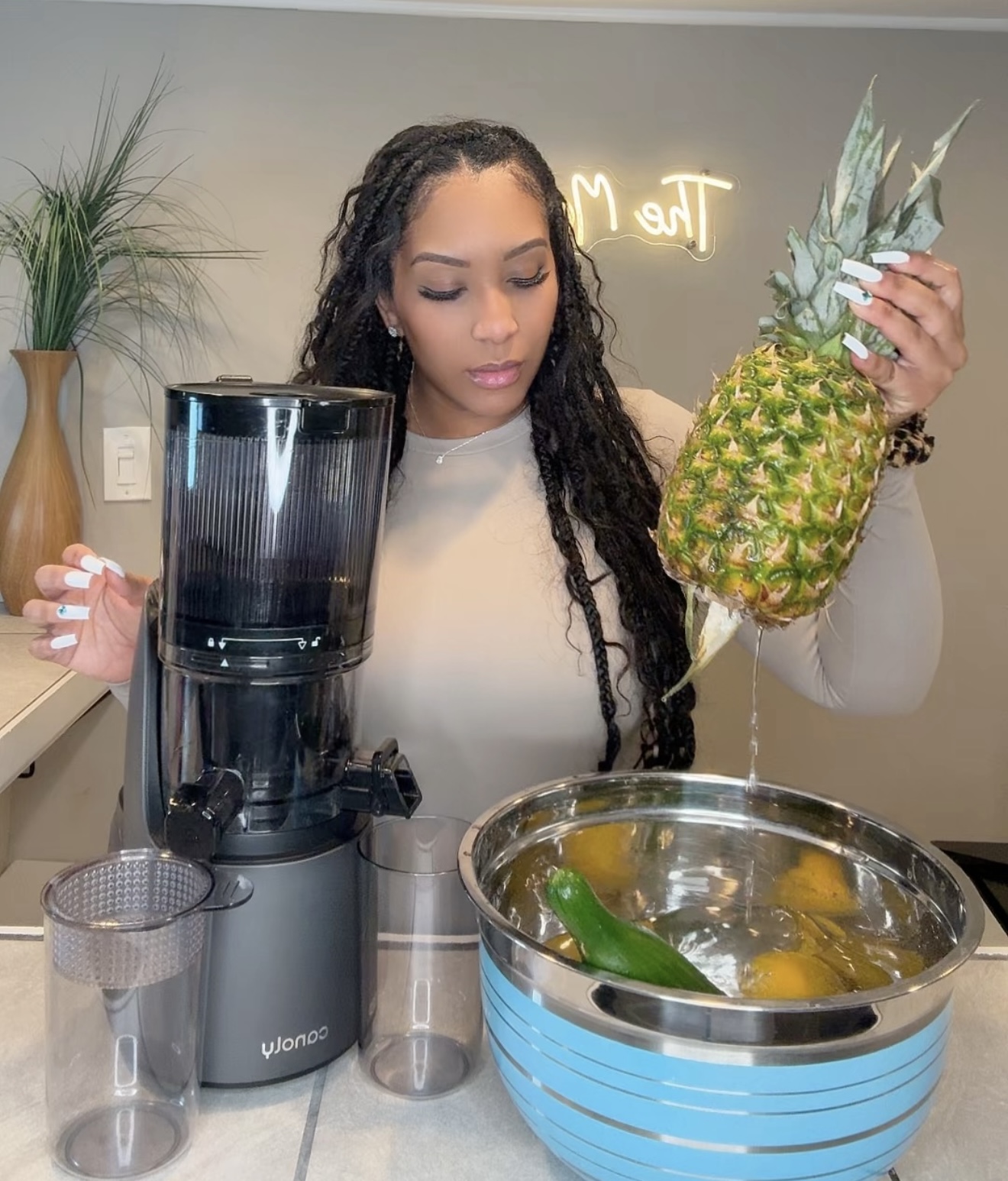 Woman holding a pineapple in a modern kitchen with a blender and a bowl of fruit, plants, and a neon sign in the background.