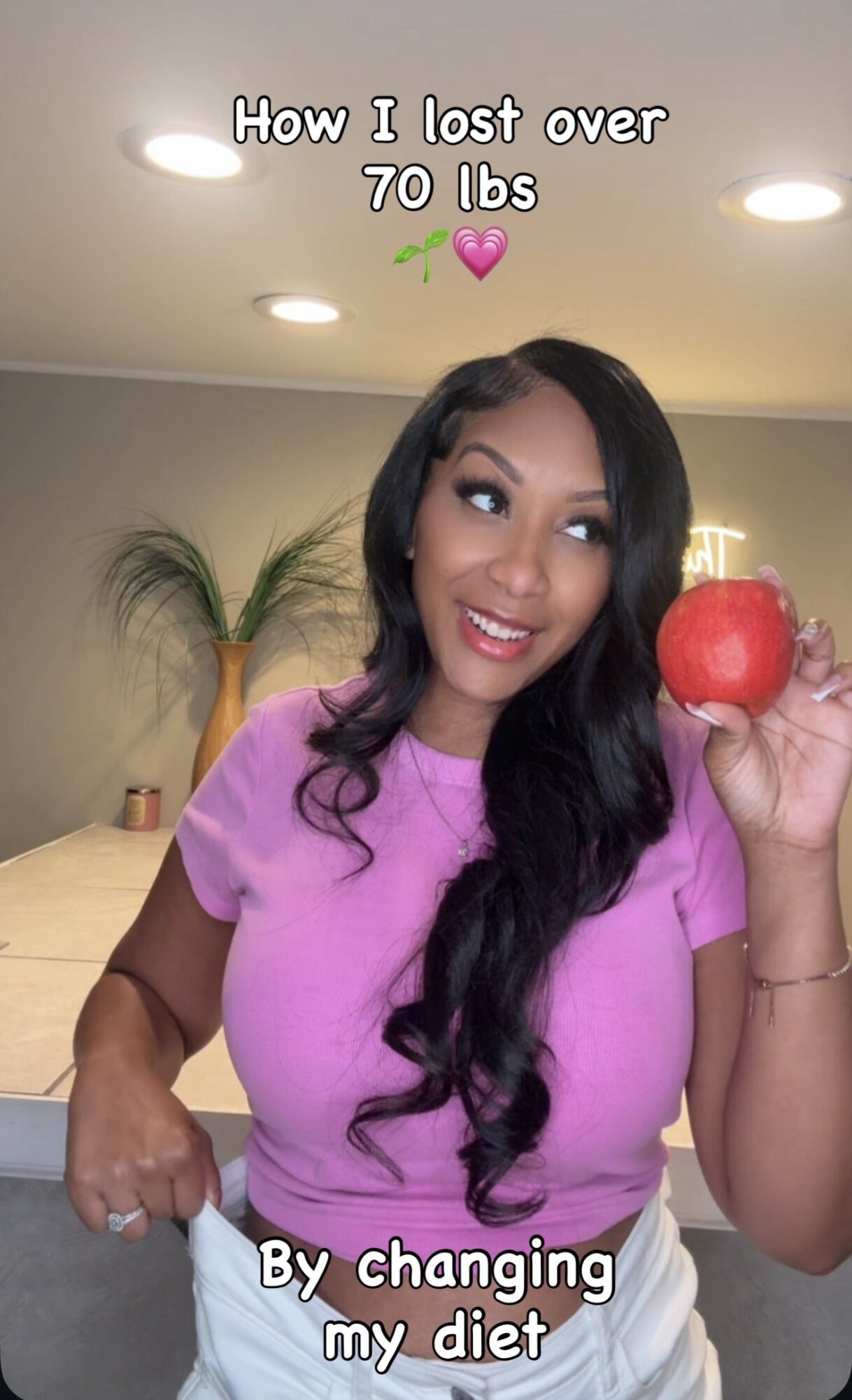 Woman with long dark hair holding a red apple, smiling in a room with a plant and ceiling lights.