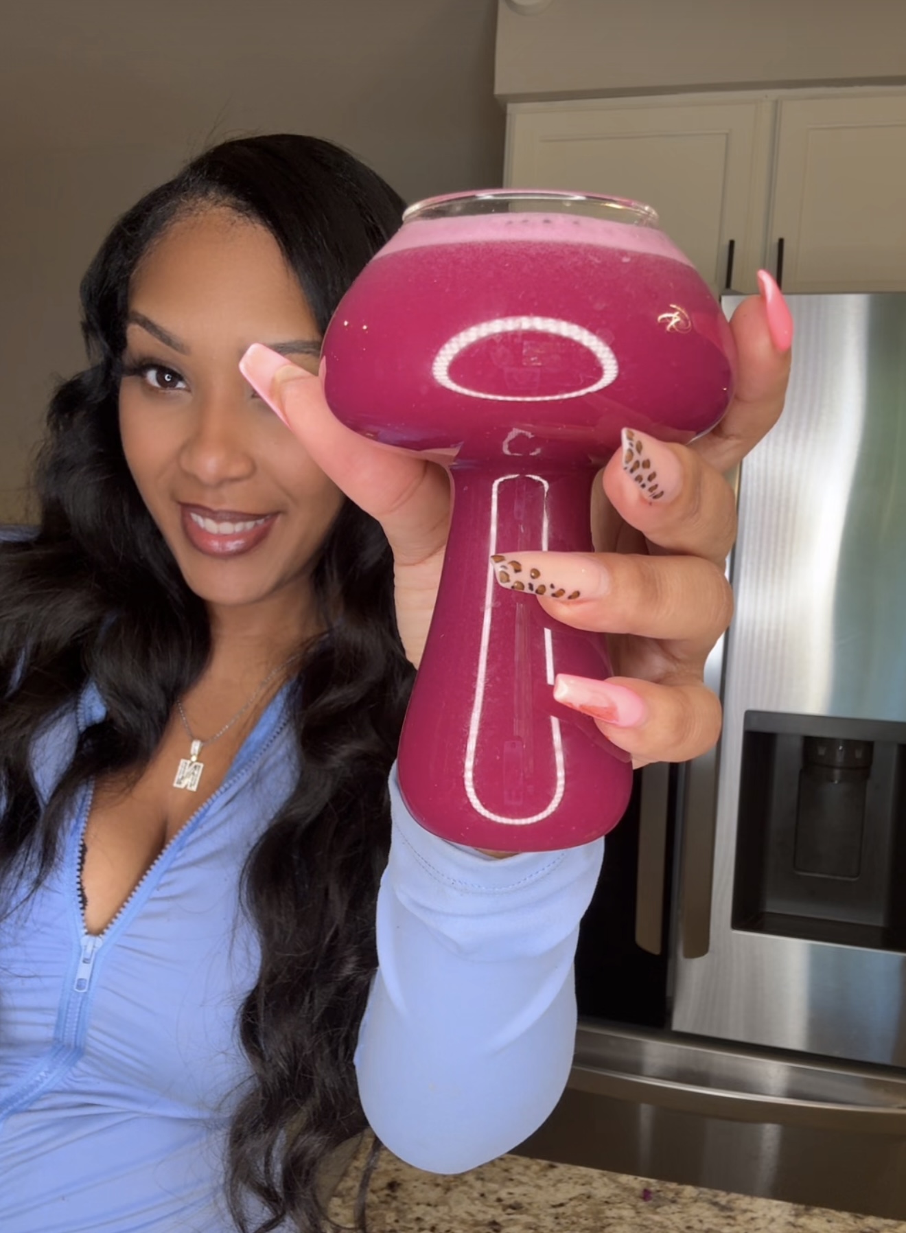 Woman holding a pink handheld device with a large bowl of pink liquid, smiling, in a kitchen setting.