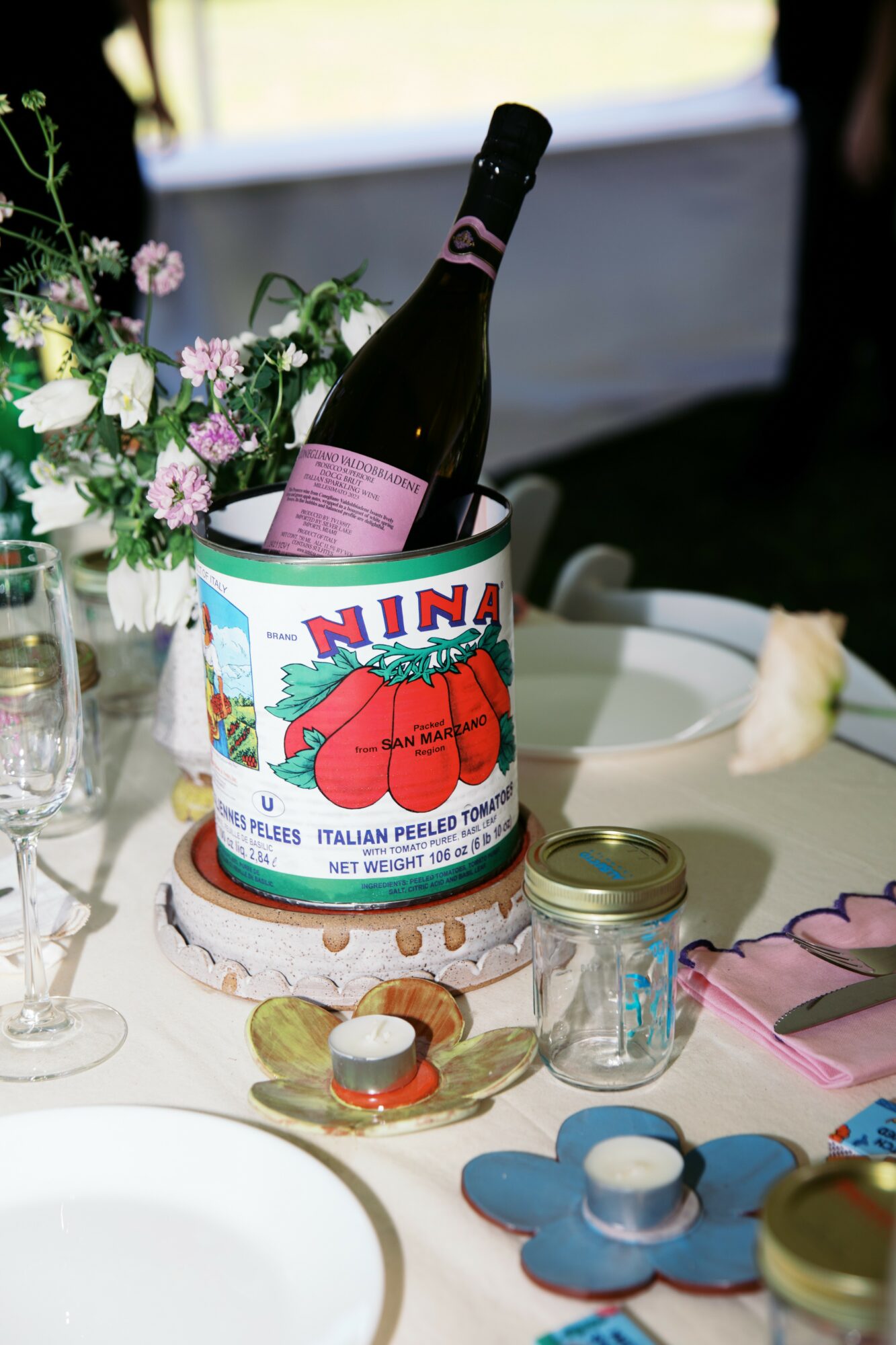 Bottle of wine in a colorful container on a table with flowers, glasses, and decorative items.