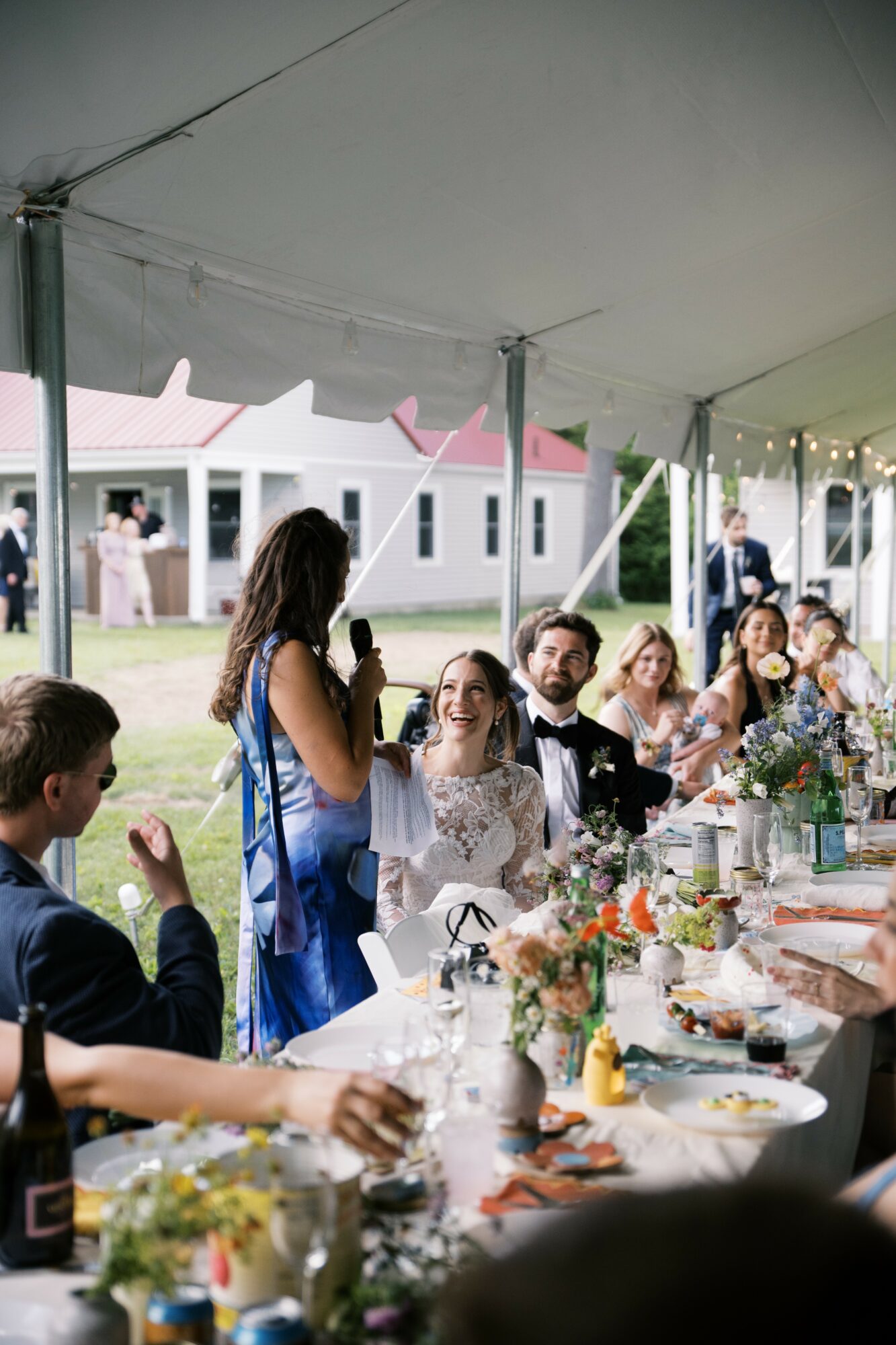 Woman speaking into microphone at outdoor wedding reception under tent, guests seated at decorated table, smiling and listening.