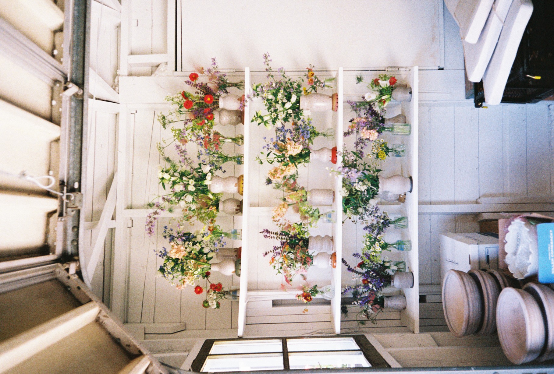 White wooden shelves with colorful flowers arranged in vases, set against a white wooden wall, viewed from above.