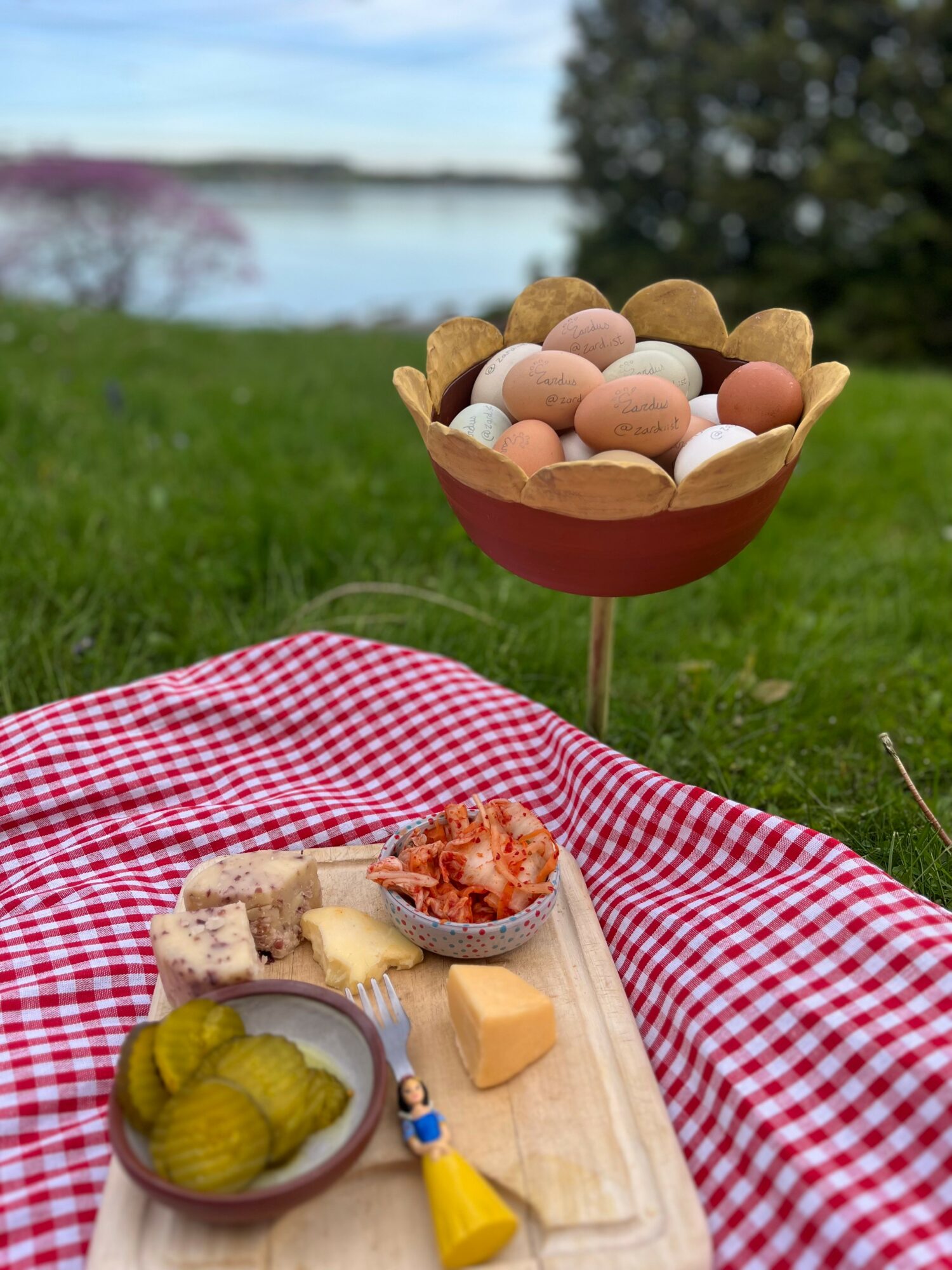 Easter eggs in a bowl on a stand, a picnic blanket with cheese, pickles, and a small toy, outdoors near water and trees.