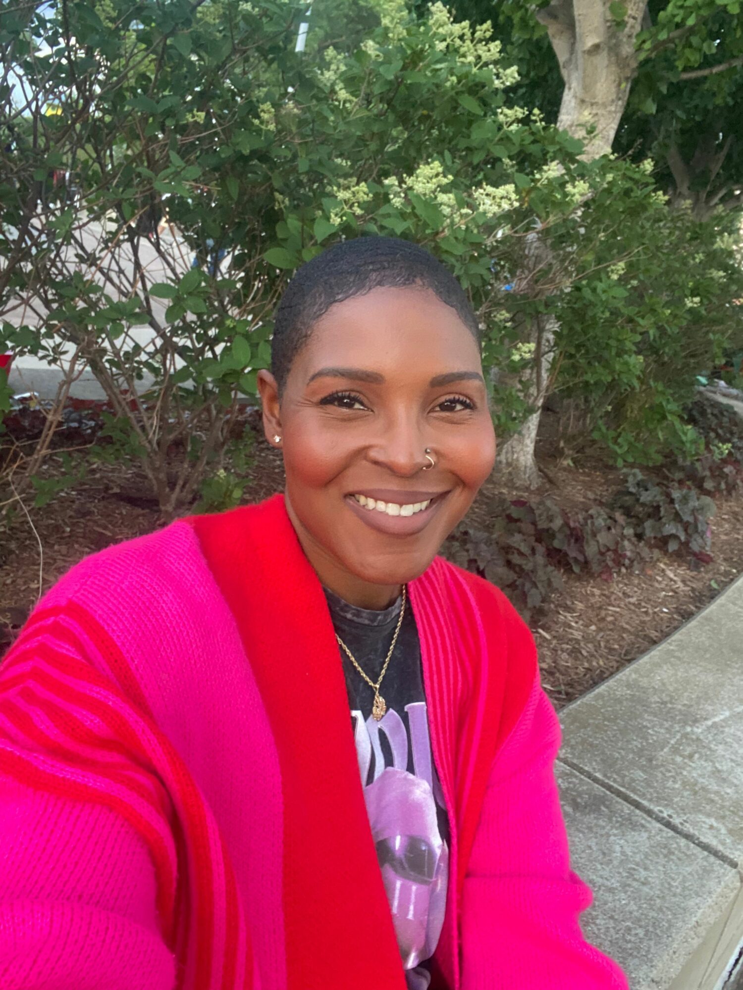Smiling woman with short hair, wearing a pink jacket and a black and white striped shirt, outdoors near greenery.