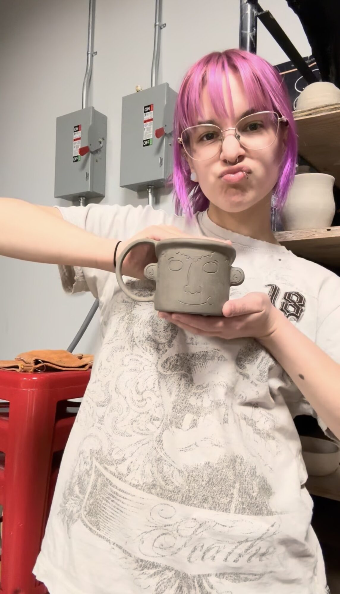 Person with pink hair and glasses holding a ceramic mug, standing in a pottery studio.