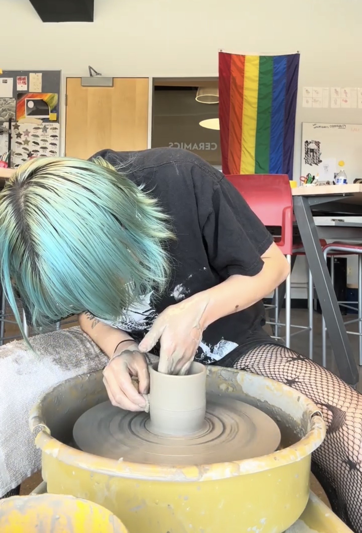 Person with colorful hair working on a pottery wheel in a room with a rainbow flag, desk, and chair.