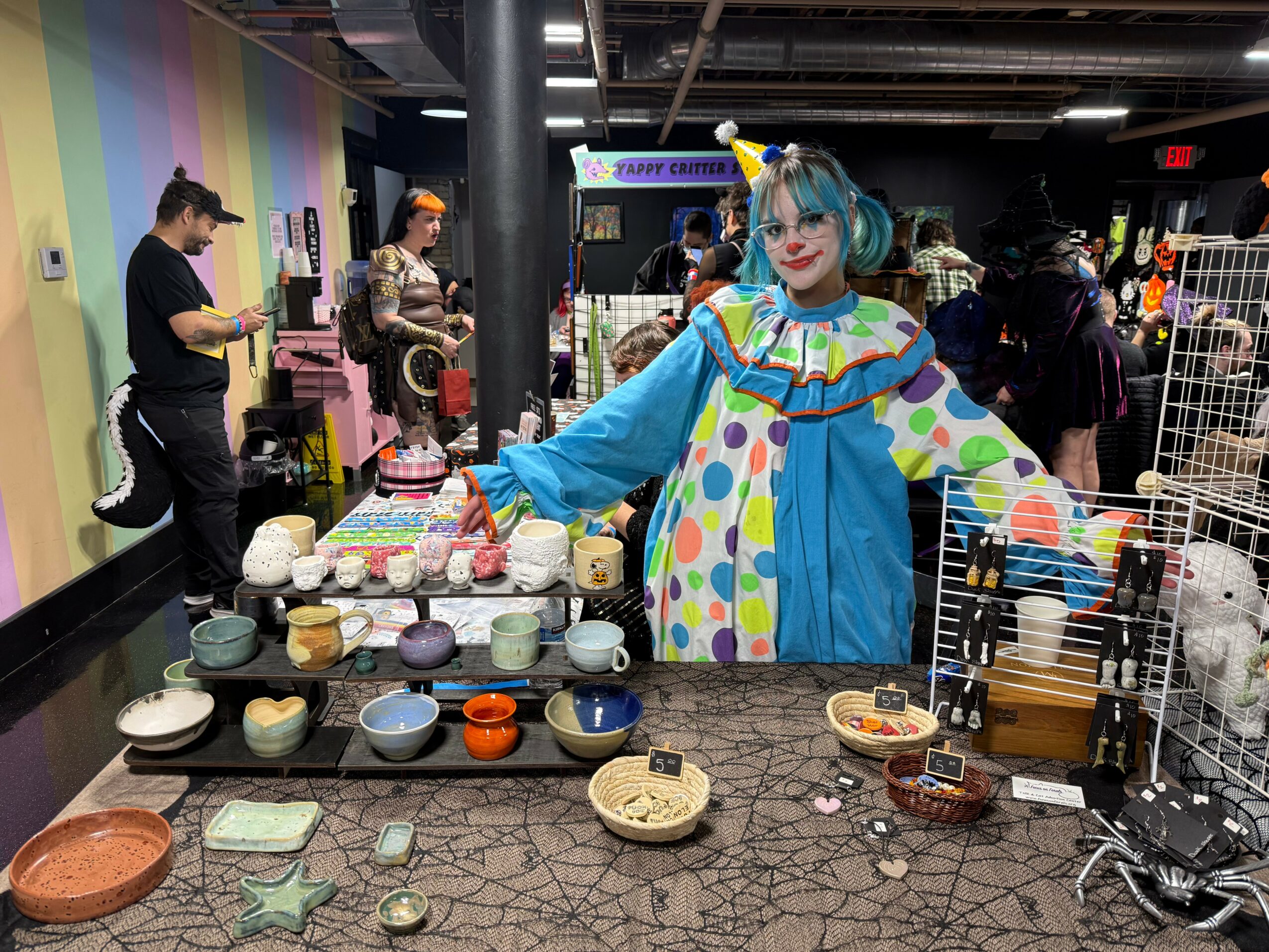 Person dressed as a clown with blue hair and colorful costume stands behind a table with various items at a market or fair, with people browsing in the background.