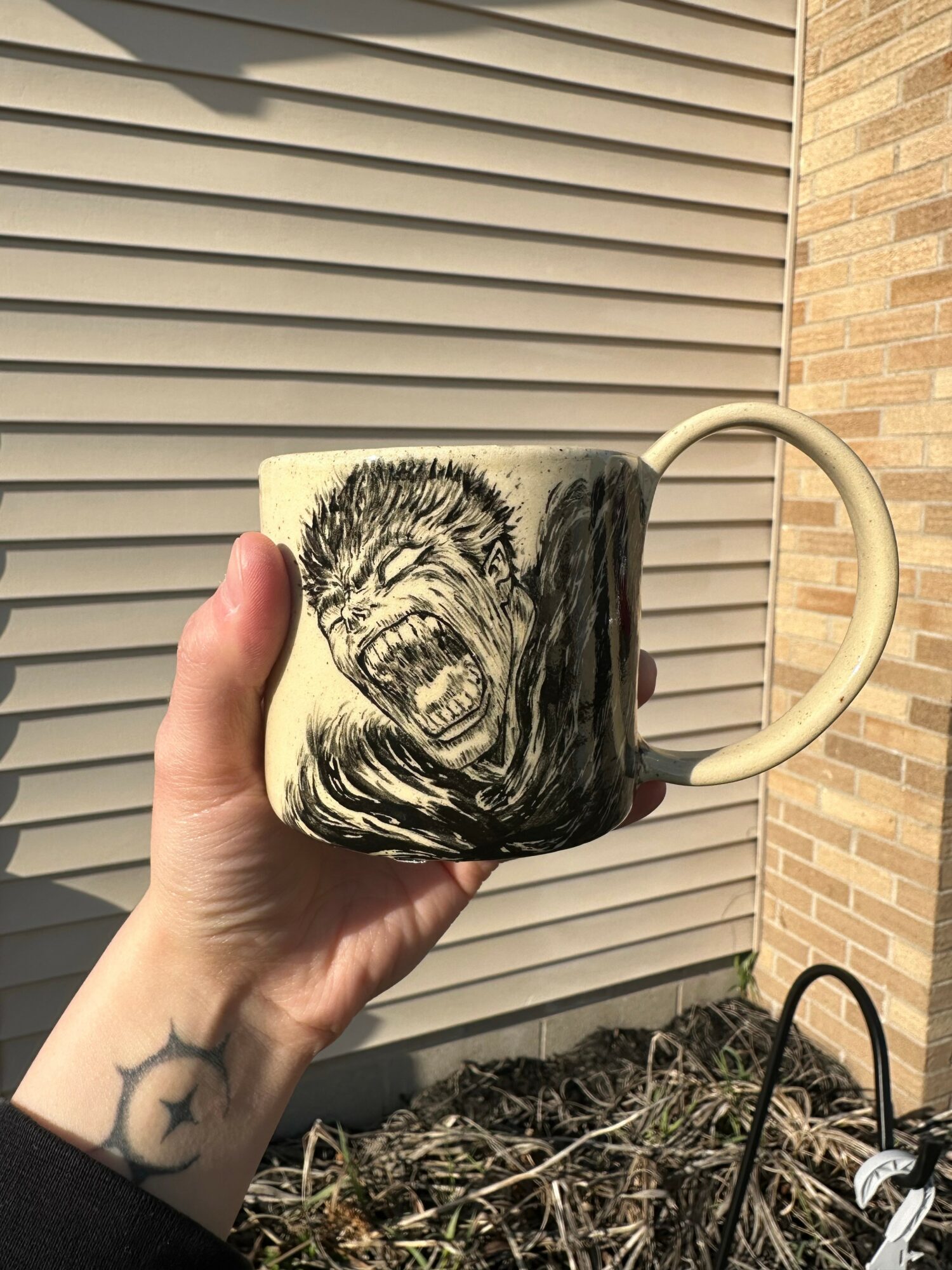 Hand holding a mug with a roaring lion illustration, background of horizontal siding and brick wall, tattoo on wrist, outdoor setting