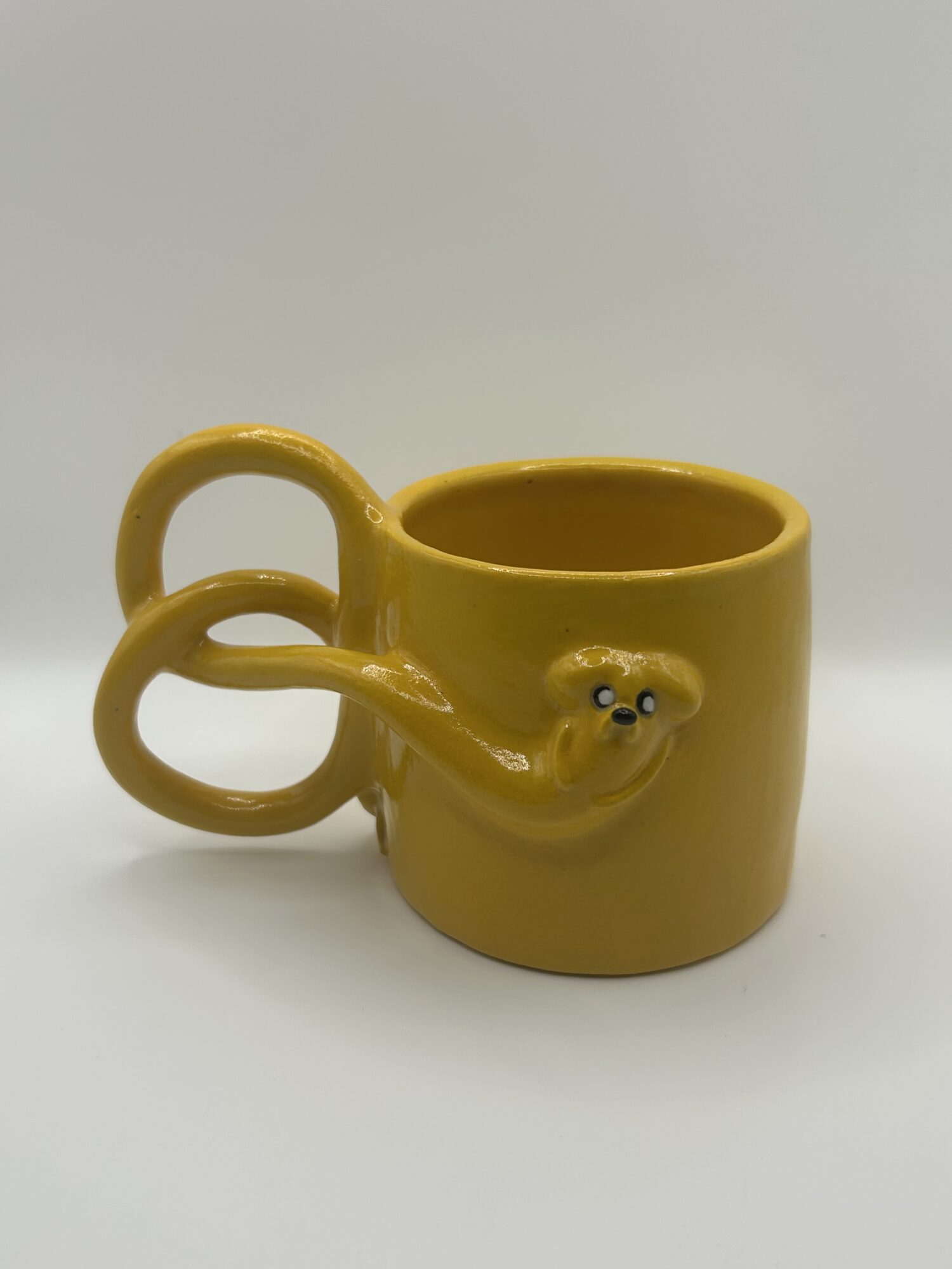 Yellow mug with a face and two handles, placed on a white surface against a white background.