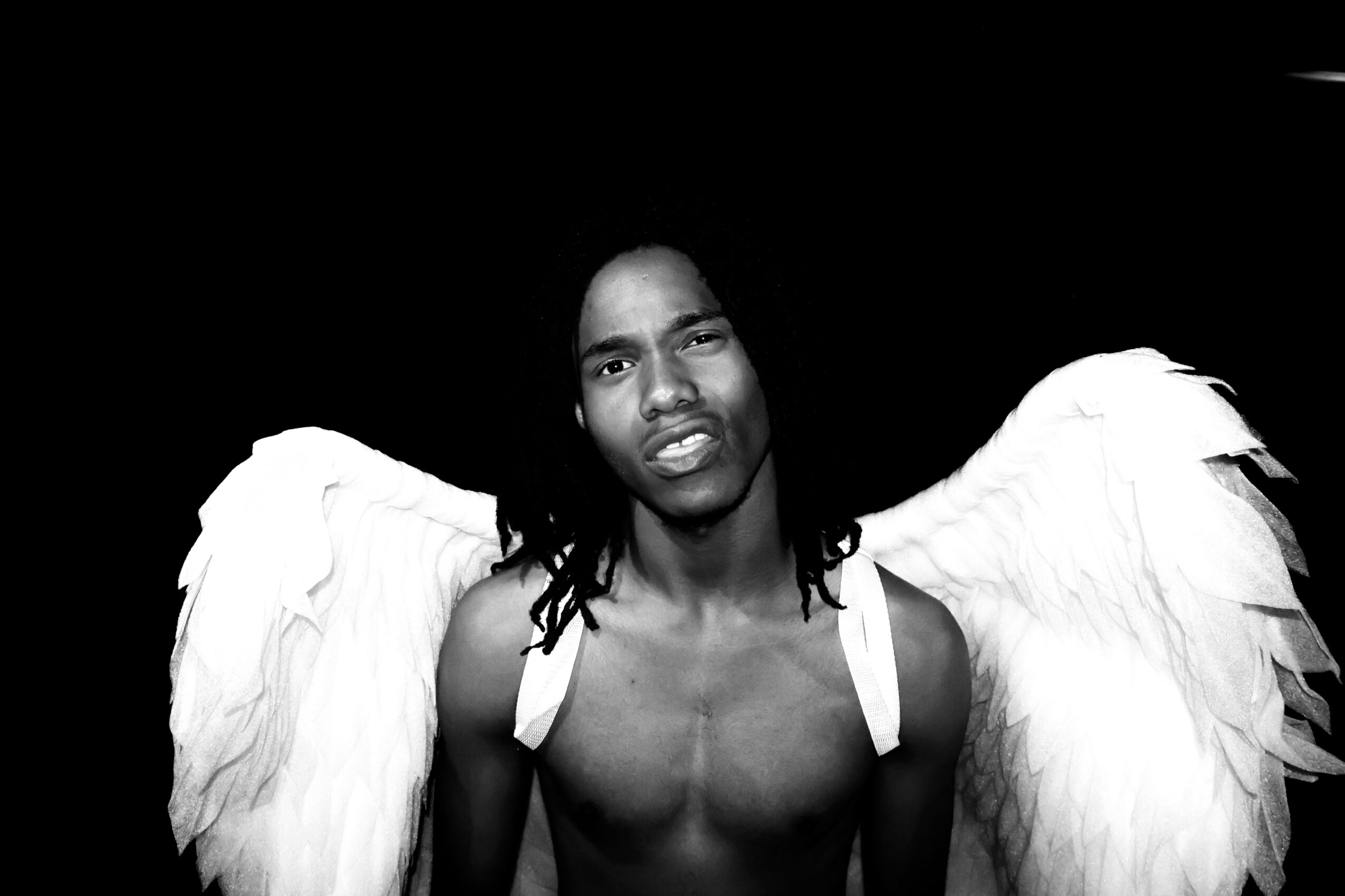 Young man with angel wings against a black background, looking at the camera, shirtless, with long dreadlocks.