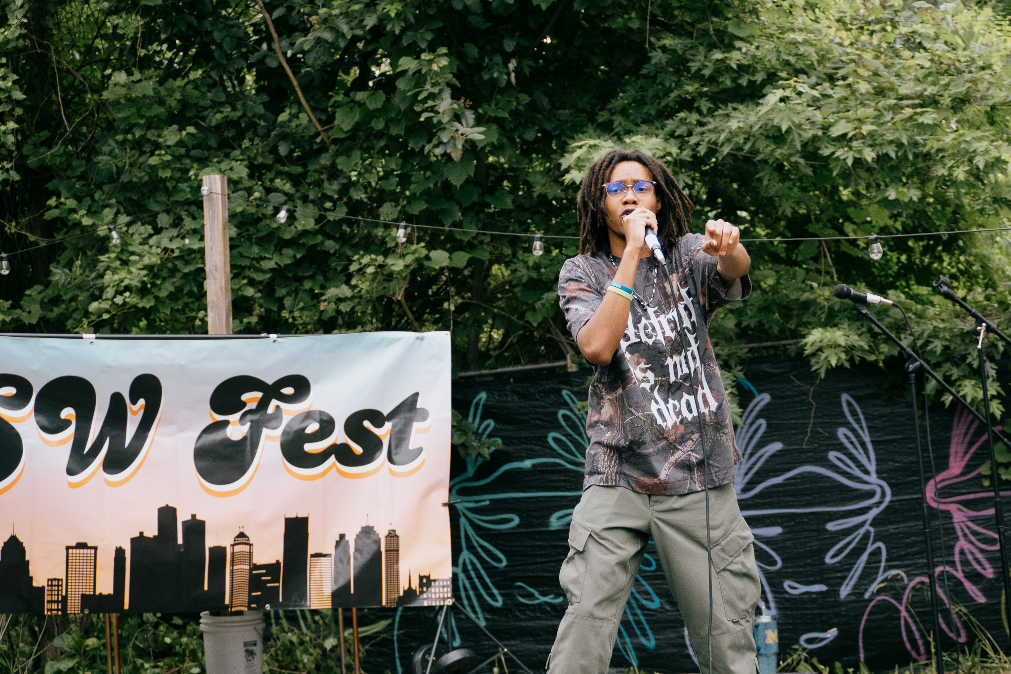 A person with long hair and sunglasses sings into a microphone on an outdoor stage with trees in the background.