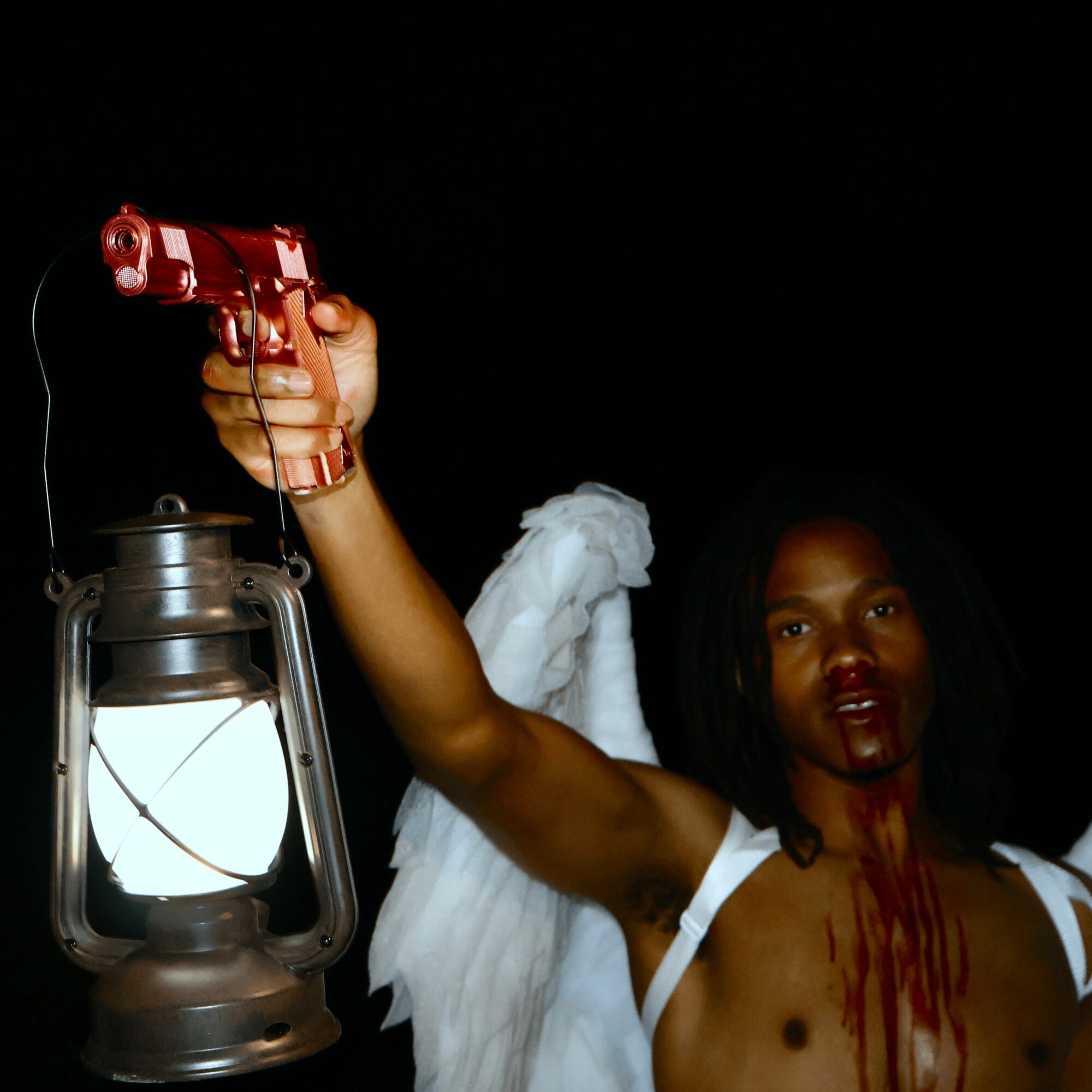 Person with dark skin and black hair holding a red toy gun, wearing a white top, with a white cloth on shoulder, against a black background.