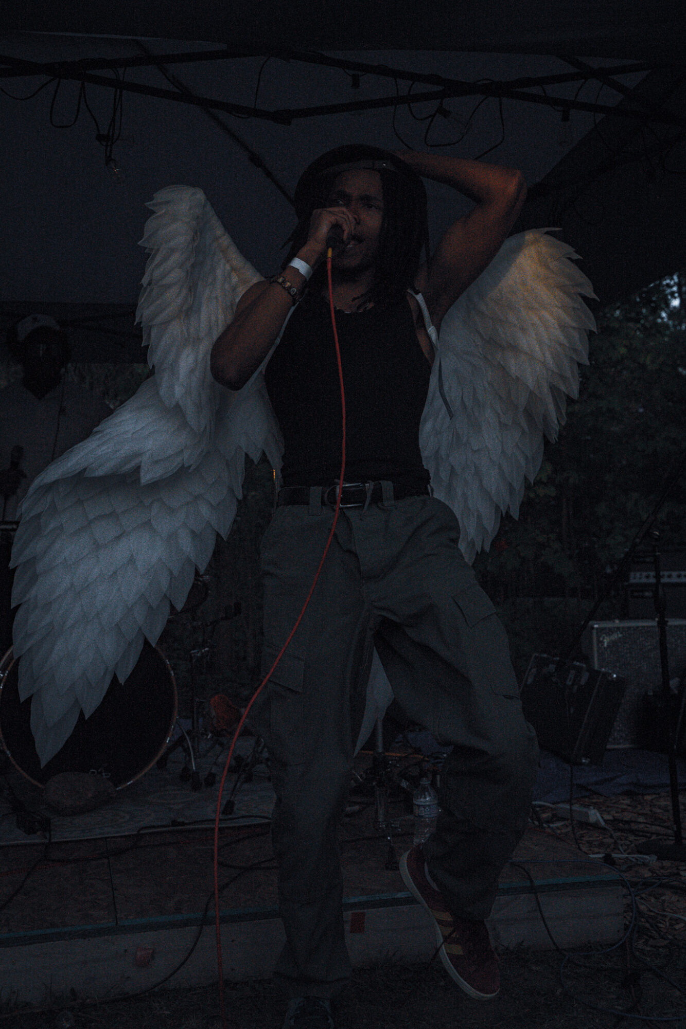 Person with large white feathered wings, holding microphone, standing on stage with dark background.