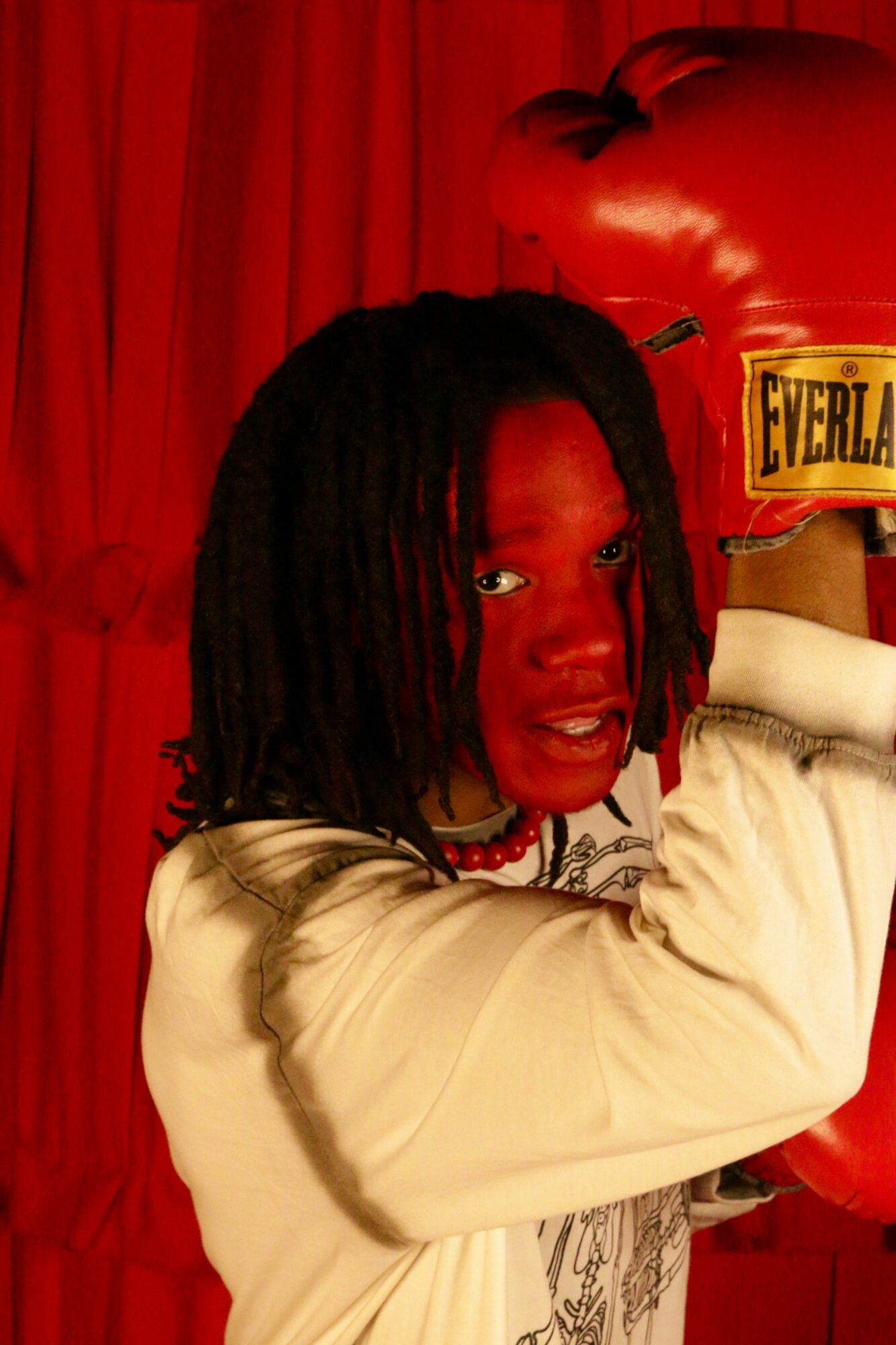 Person with dreadlocks wearing a beige jacket, holding a red boxing glove, in front of a red curtain.