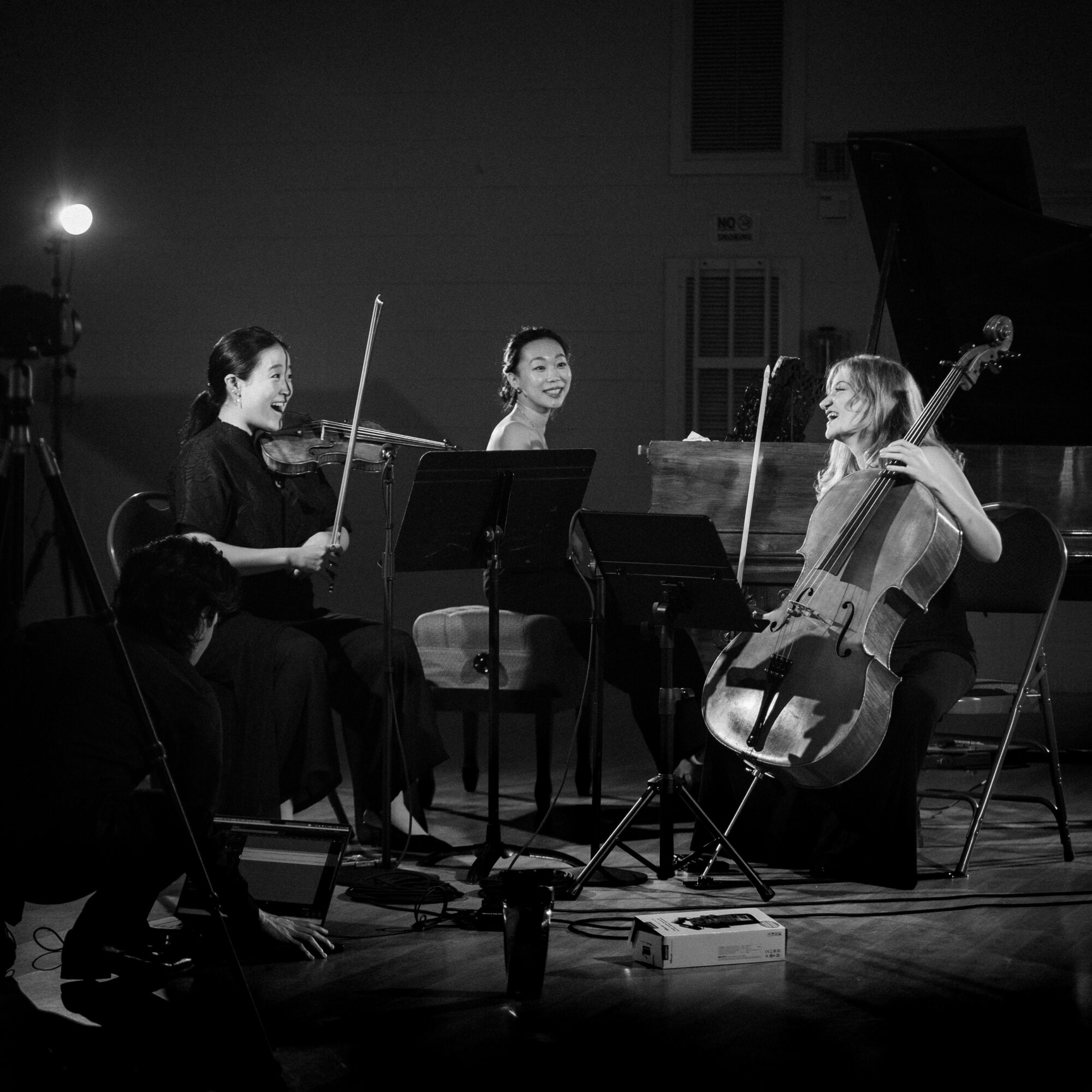 Four musicians perform on stage, three playing violins and one playing a cello, in a black-and-white setting.